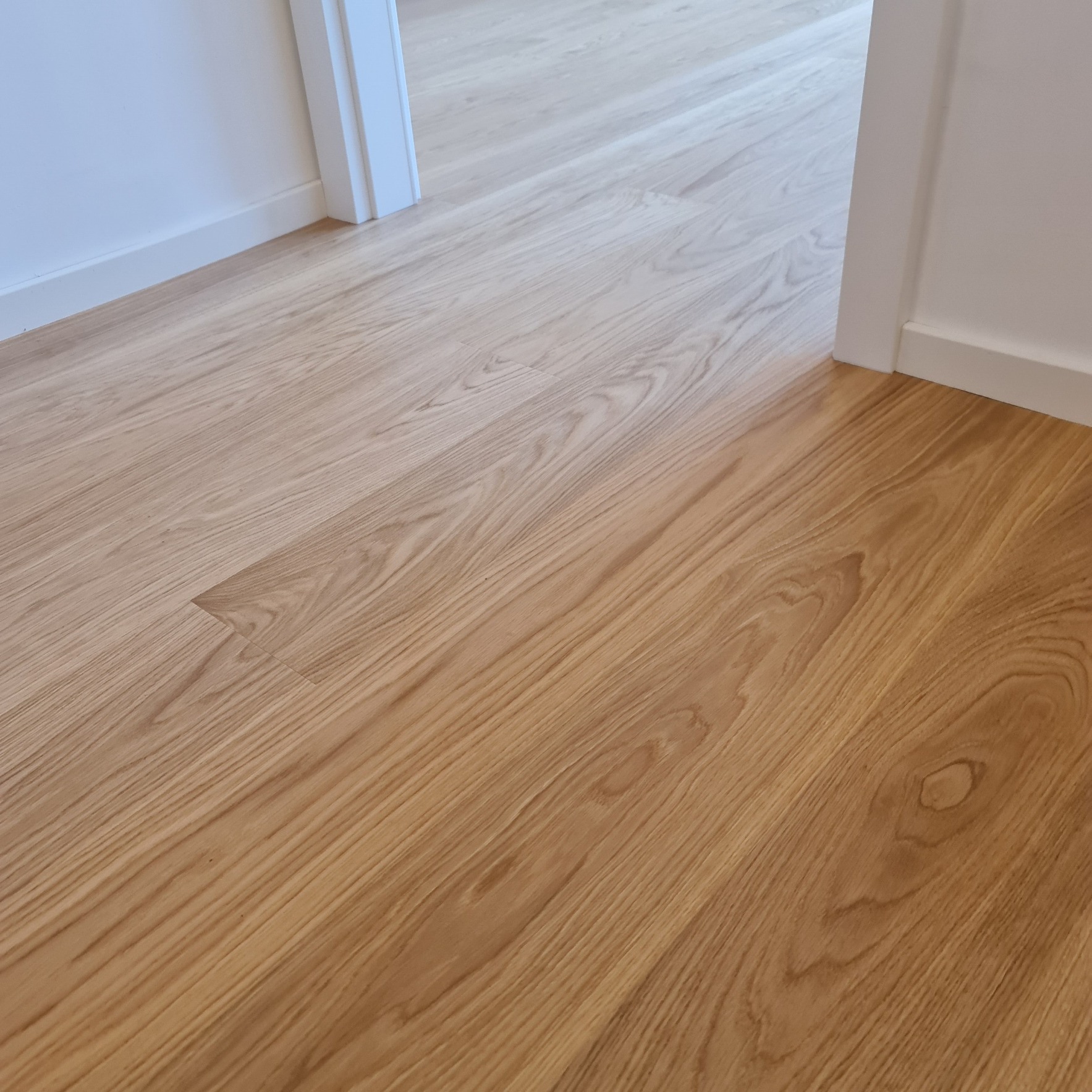 Jasny parkiet ułożony wzdłuż korytarza, widoczny fragment białej ściany i futryny drzwi. Naturalne oświetlenie podkreśla strukturę drewna.