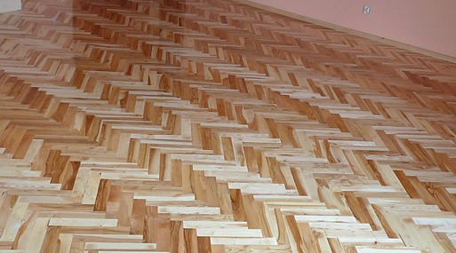 Jasny parkiet ułożony we wzór jodełki na podłodze, widok z góry pod kątem. Naturalne drewno z widocznymi słojami. Fragment ściany w tle.