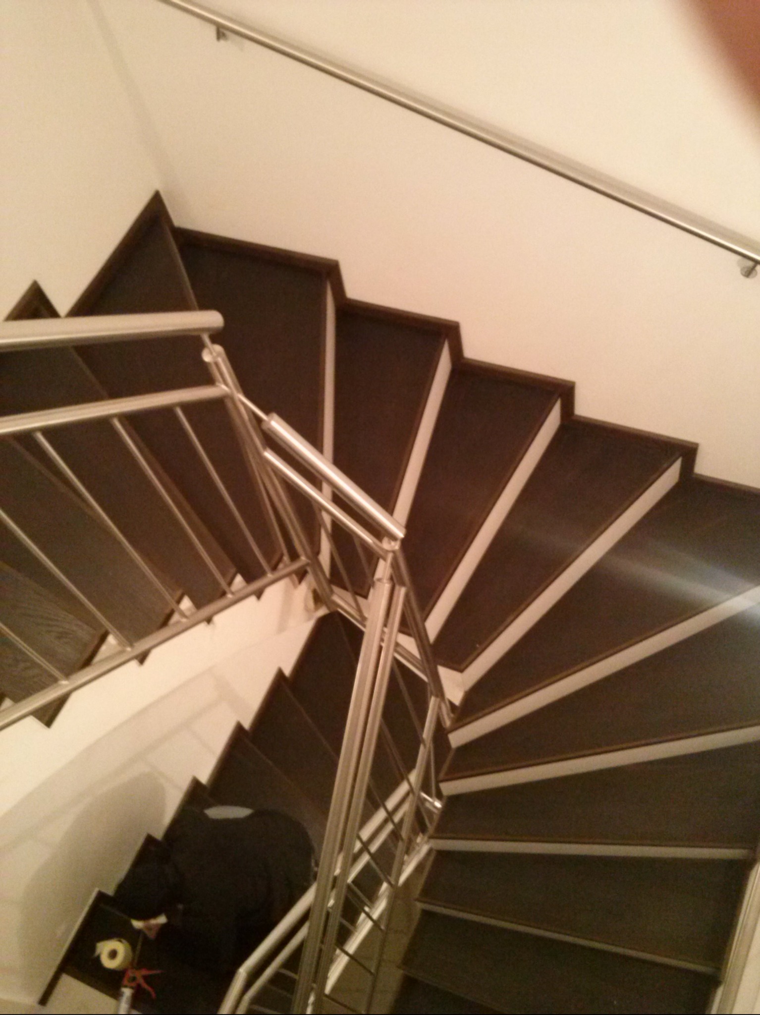 Spiralne schody z ciemnego drewna z metalową balustradą, widok z góry. Kontrastowe białe krawędzie stopni. Na dole widoczna osoba w ciemnym ubraniu.