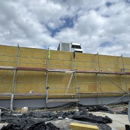 E.B.Bud Ewa Pytel - Ocieplanie budynku wełną mineralną na rusztowaniu. Widoczne materiały budowlane i wentylacja na dachu. Prace elewacyjne w toku, z bliska, w dzień.