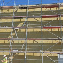 E.B.Bud Ewa Pytel - Ocieplanie elewacji budynku wełną mineralną, widoczne rusztowania i pracownik w kamizelce odblaskowej. Żółta izolacja termiczna na ścianie.