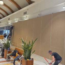 E.B.Bud Ewa Pytel - Montaż ścianki działowej z płyt kartonowo-gipsowych w przestrzeni handlowej. Pracownicy na drabinie i przy podłodze, widoczne narzędzia i materiały budowlane.