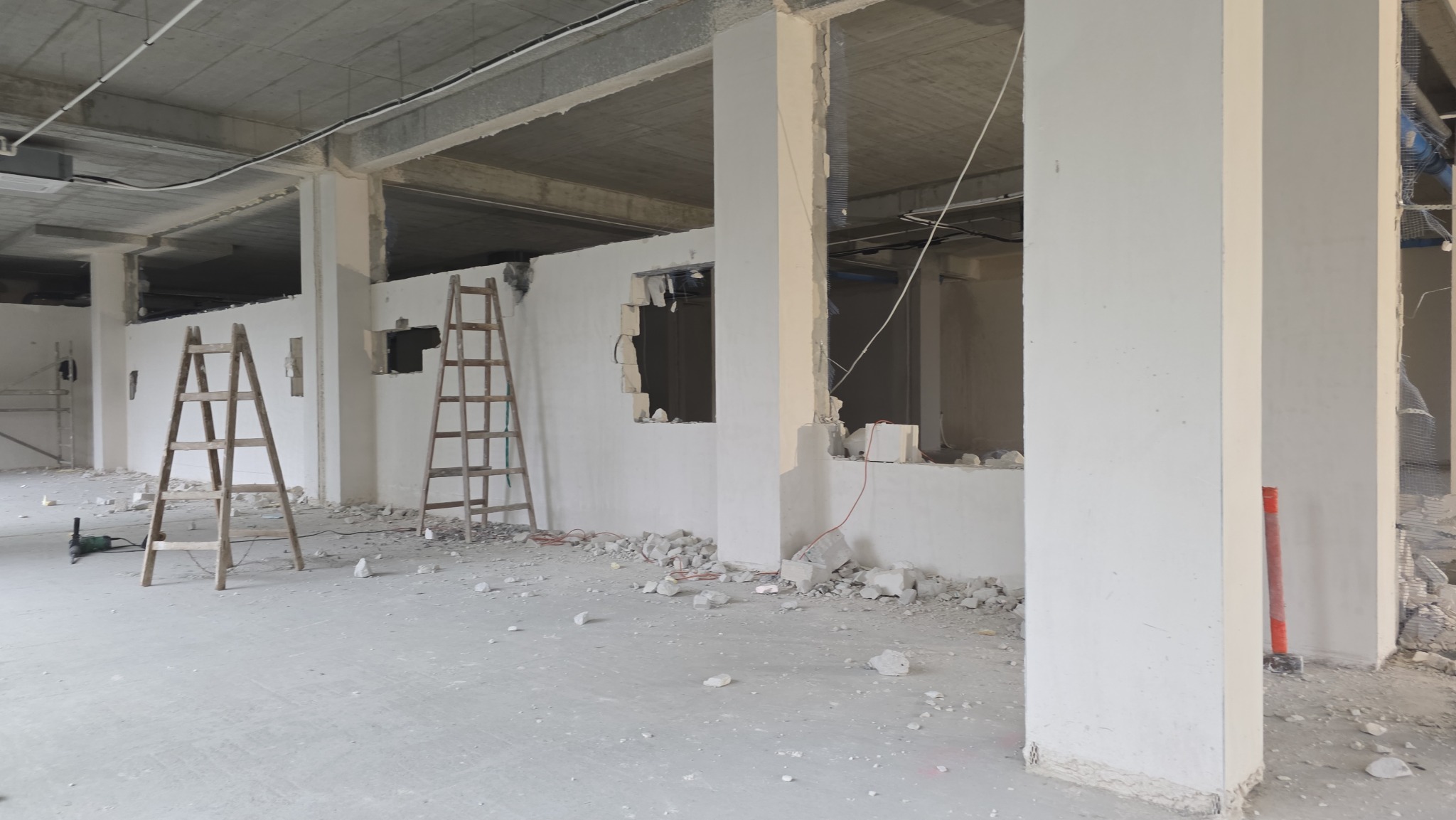 Wnętrze w trakcie rozbiórki: fragment ściany z otworami, gruz na podłodze, dwie drewniane drabiny. Widoczne betonowe filary i sufit. Surowy stan, przygotowanie do dalszych prac.