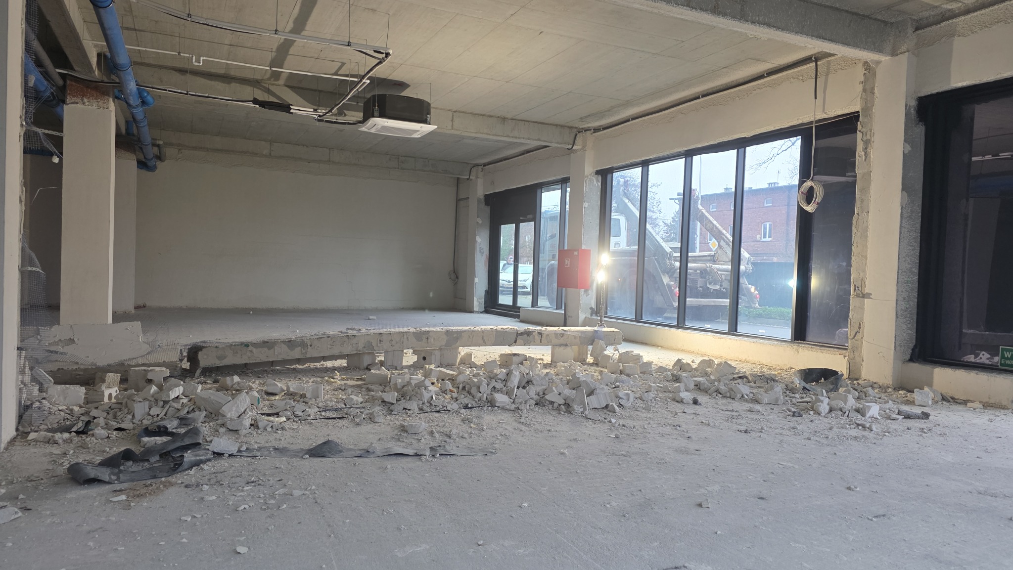 Wnętrze w trakcie rozbiórki: gruz na podłodze, odsłonięty beton, widoczne instalacje pod sufitem. Przez okna widać budynek i pojazd budowlany.
