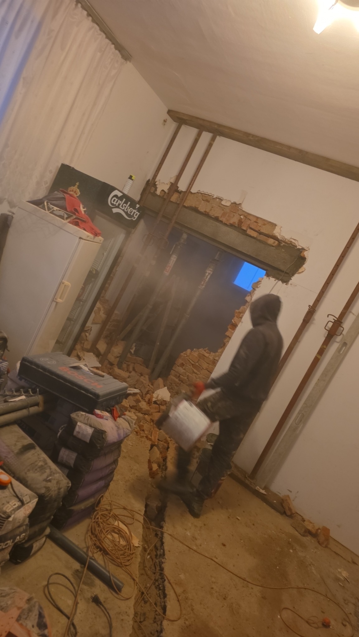 Demontaż ściany z cegły w trakcie realizacji. Widoczne gruz, narzędzia i pracownik w odzieży roboczej. Podpory wzmacniające konstrukcję. Widok na zewnątrz przez otwór.