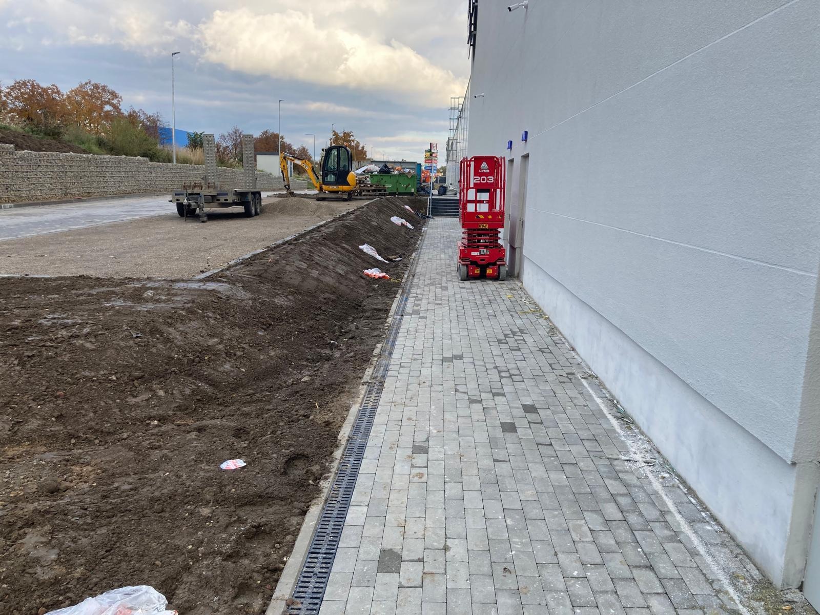 Przygotowanie terenu pod budowę: wykop, drenaż, kostka brukowa, podnośnik koszowy, mini koparka. Widok na elewację budynku i niebo z chmurami.