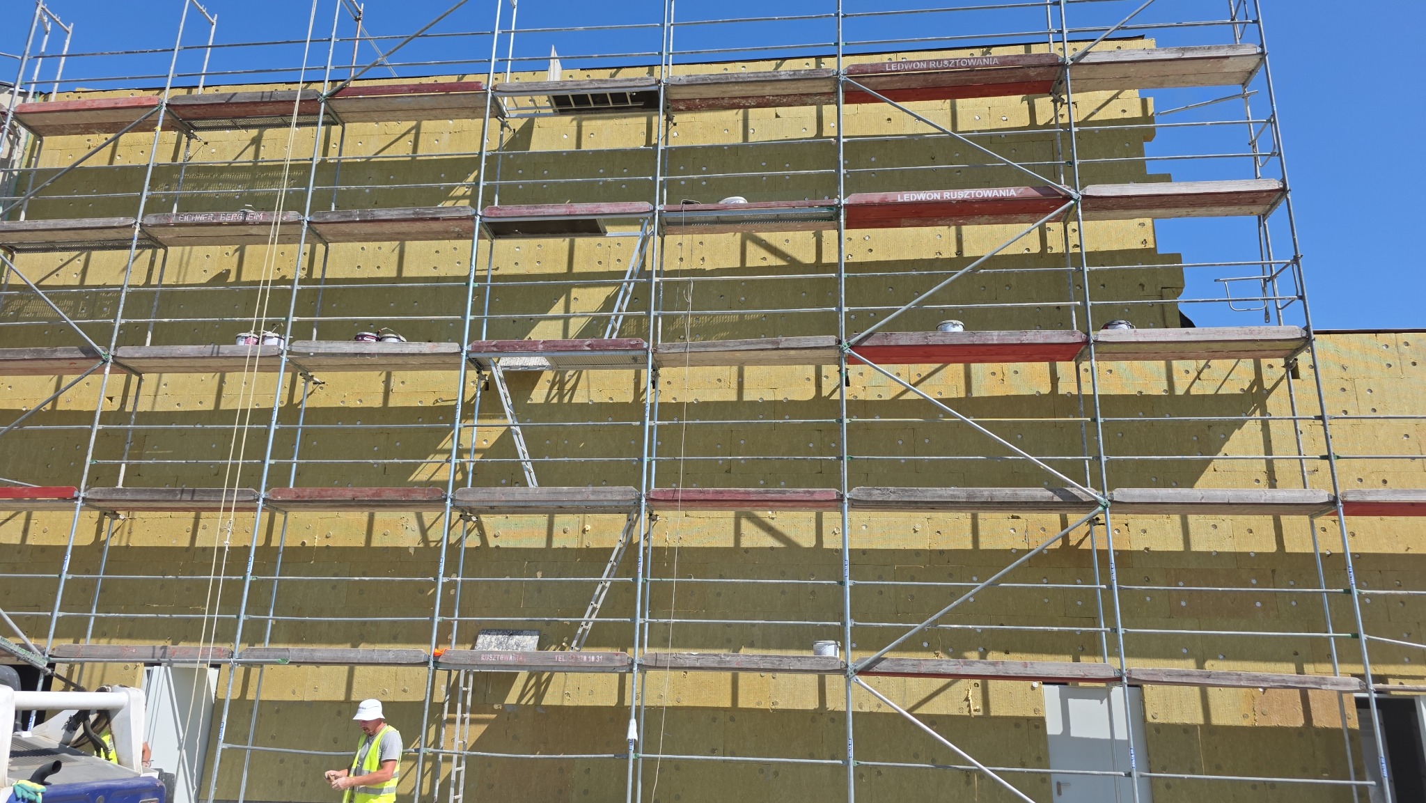 Ocieplanie elewacji budynku wełną mineralną, widoczne rusztowania i pracownik w kamizelce odblaskowej. Żółta izolacja termiczna na ścianie.