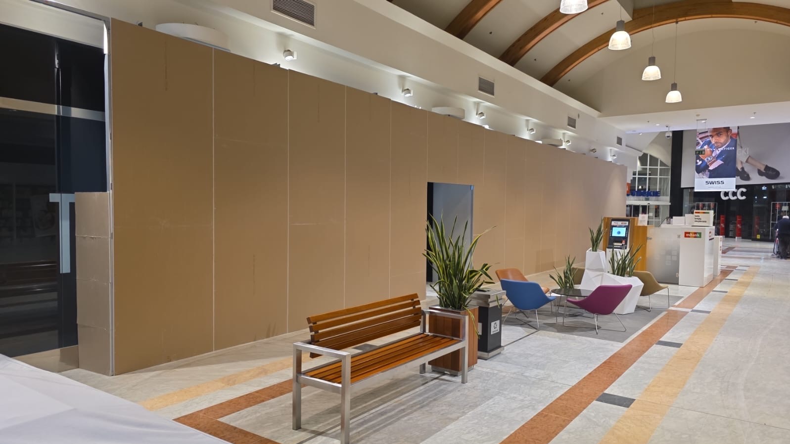 Tymczasowa ścianka działowa z płyty w centrum handlowym, z ławką i roślinami doniczkowymi na pierwszym planie. Widoczne elementy infrastruktury centrum.