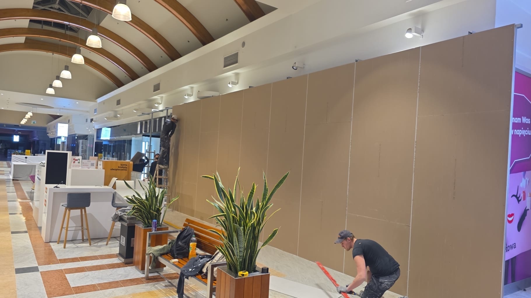 Montaż ścianki działowej z płyt kartonowo-gipsowych w przestrzeni handlowej. Pracownicy na drabinie i przy podłodze, widoczne narzędzia i materiały budowlane.