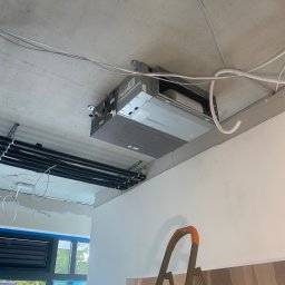 Airfun WŁODZIMIERZ MELNAROWICZ - Sufitowa jednostka klimatyzacyjna w trakcie montażu, widoczne rury i przewody. W tle okno i drabina. Surowy stan wykończenia wnętrza.