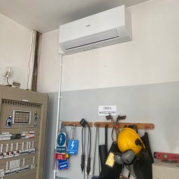 Airfun WŁODZIMIERZ MELNAROWICZ - Biały klimatyzator Haier zamontowany na ścianie w pomieszczeniu technicznym. Widoczne narzędzia i tabliczki ostrzegawcze. Oświetlenie sufitowe.