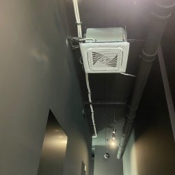 Airfun WŁODZIMIERZ MELNAROWICZ - Kasetonowa klimatyzacja Haier zamontowana na suficie w nowoczesnym biurze z szarymi ścianami i widocznymi rurami wentylacyjnymi. Oświetlenie punktowe.
