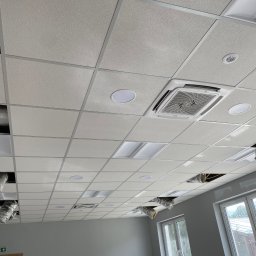 Airfun WŁODZIMIERZ MELNAROWICZ - Sufit podwieszany z zamontowaną kasetą klimatyzacyjną Haier i oświetleniem LED. Widoczne otwory na instalacje wentylacyjne i okna z widokiem na zewnątrz.
