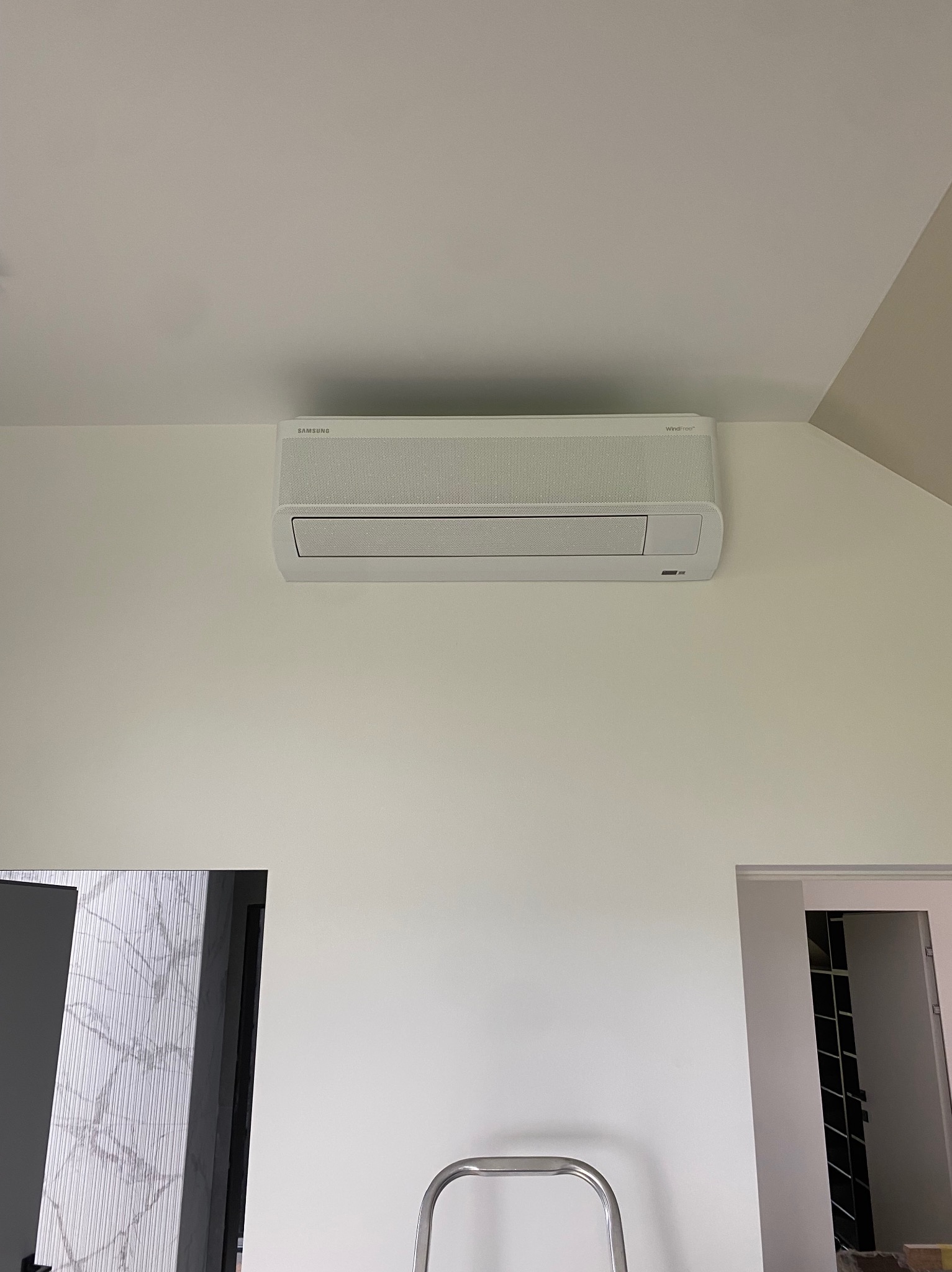 Biały klimatyzator Samsung WindFree zamontowany na ścianie w jasnym pomieszczeniu, widoczny fragment drabiny i drzwi.