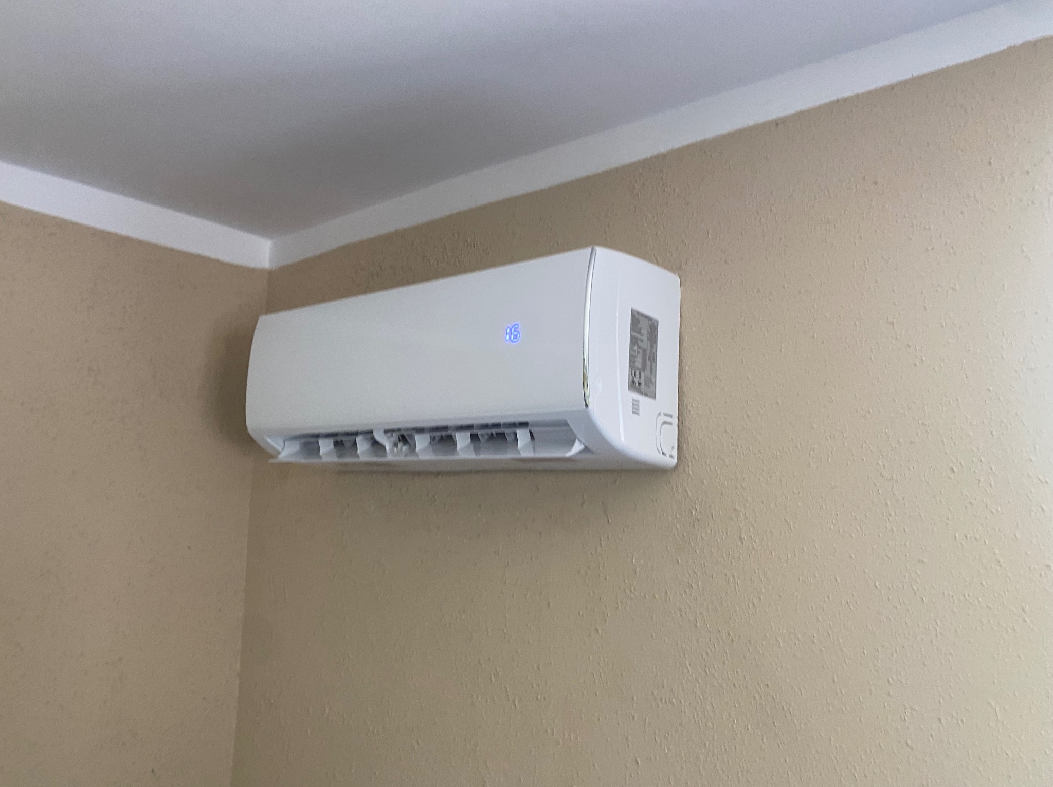 Biała klimatyzacja ścienna z wyświetlaczem temperatury, zamontowana na beżowej ścianie w rogu pomieszczenia, widok z boku.