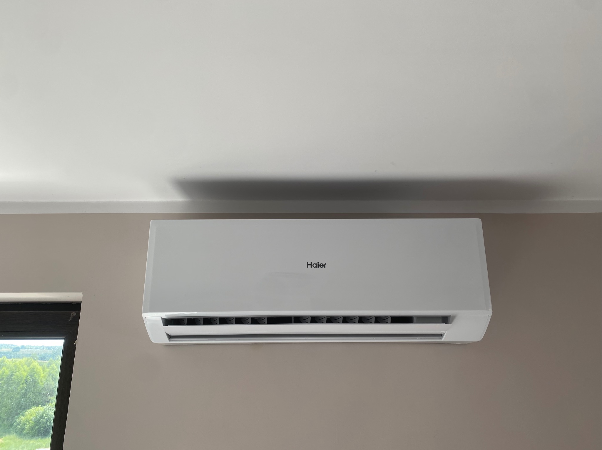Biały klimatyzator Haier zamontowany na beżowej ścianie w pomieszczeniu z widokiem na zieleń za oknem. Widoczny fragment sufitu.