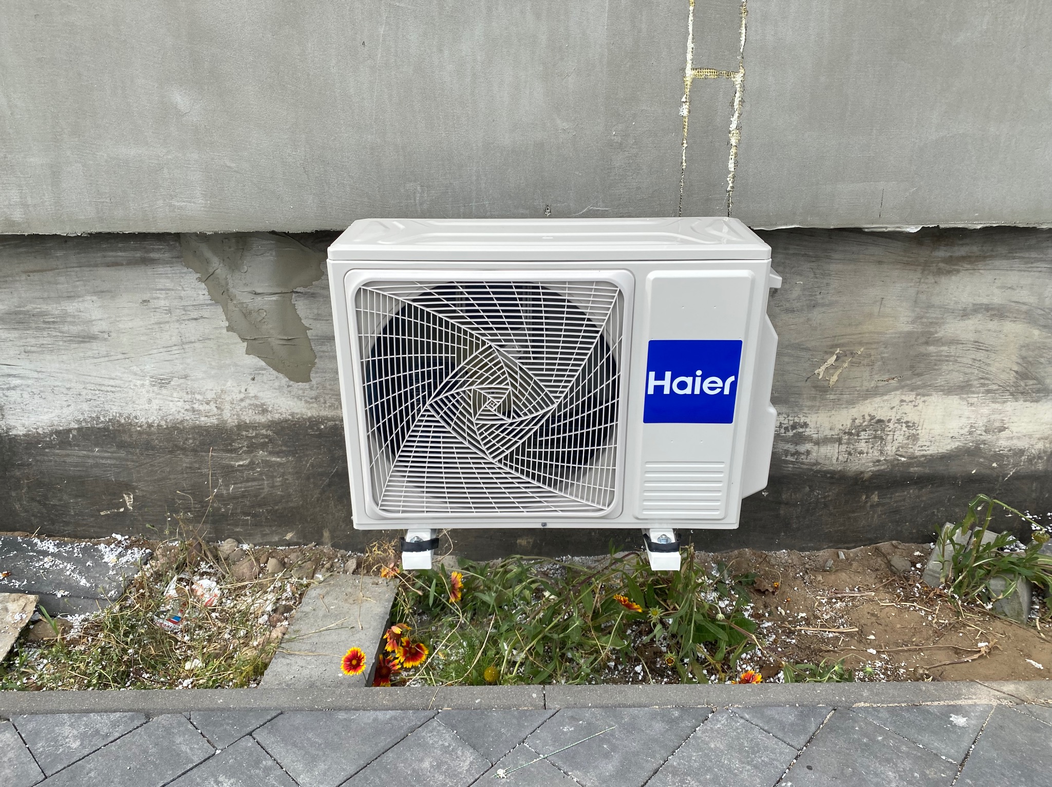 Biały klimatyzator Haier zamontowany na szarej ścianie budynku, z widocznymi elementami instalacji i roślinnością u podstawy. Ujęcie z poziomu chodnika.