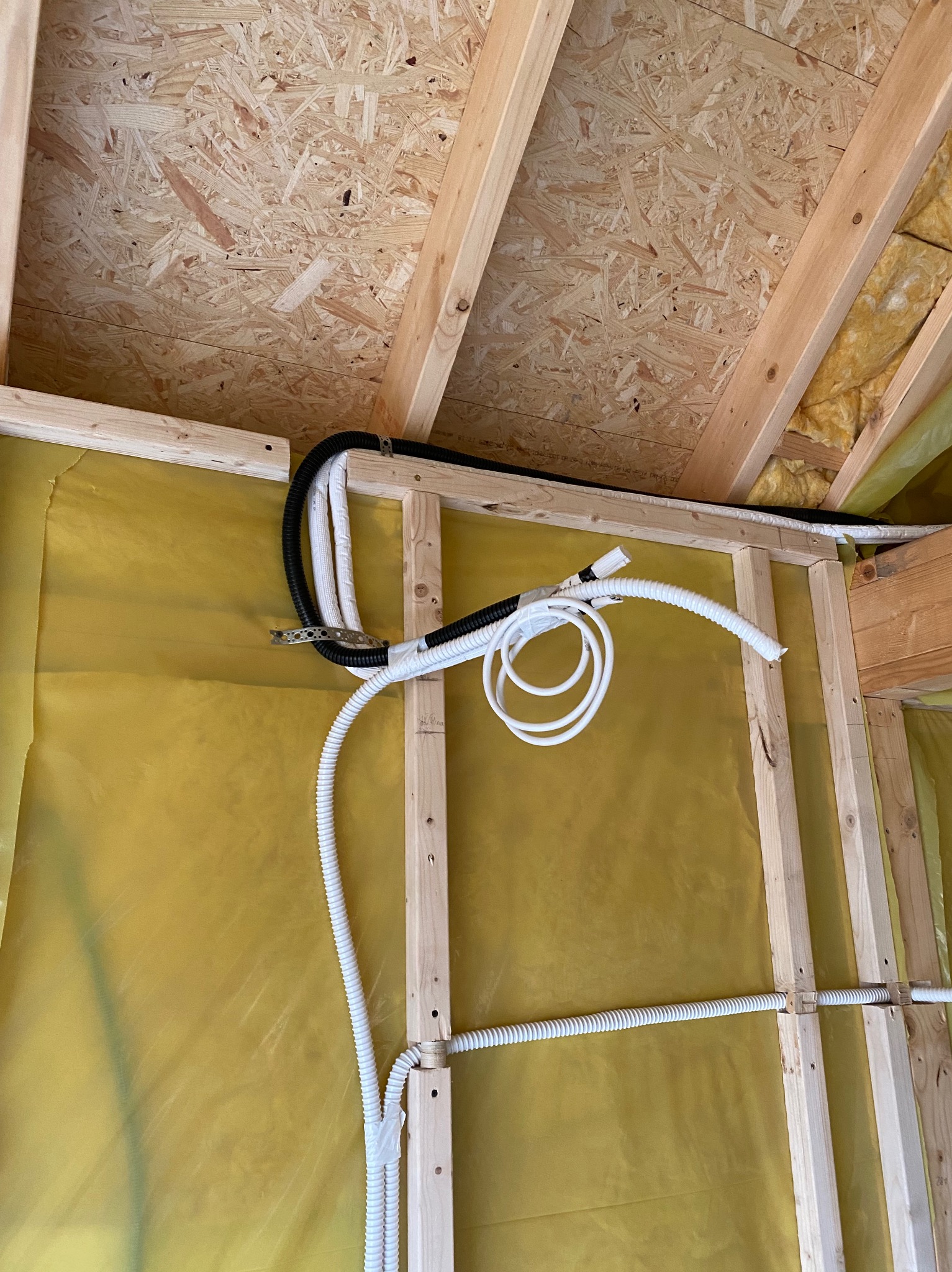 Montaż instalacji: białe i czarne rury prowadzone wzdłuż drewnianej konstrukcji ściany z żółtą folią izolacyjną. Widok z dołu na sufit z płyt OSB.