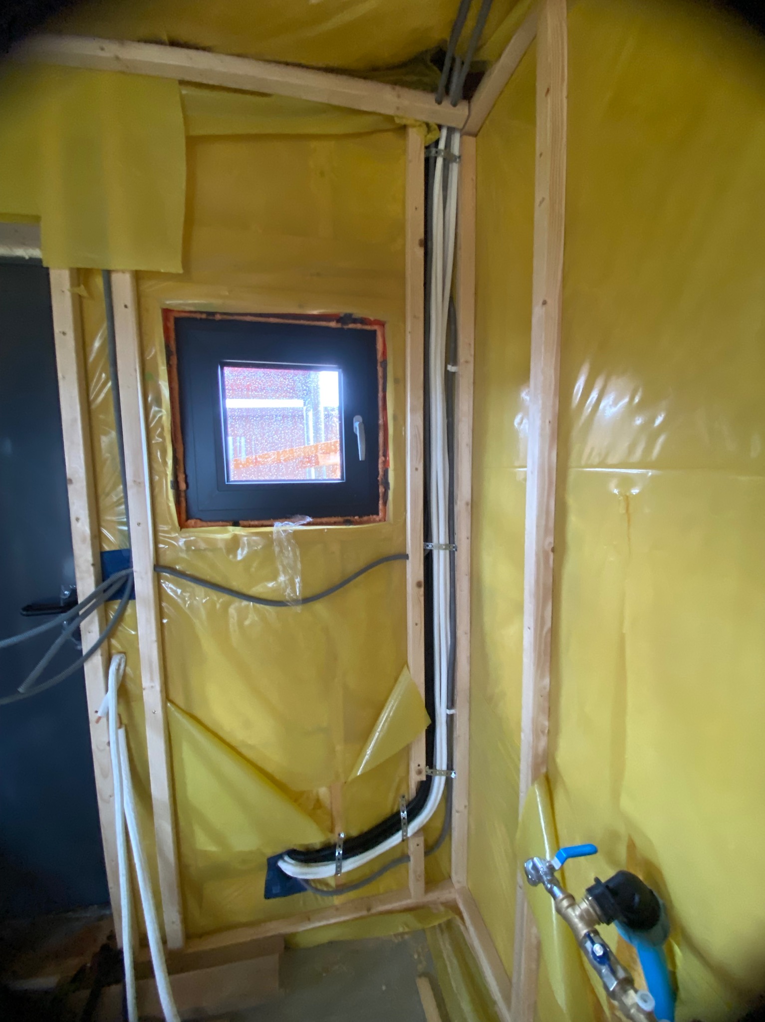 Wnętrze w trakcie prac instalacyjnych: okno, drewniana konstrukcja, żółta folia, rury klimatyzacji i instalacji wodnej. Widoczne elementy montażowe i przyłącze.
