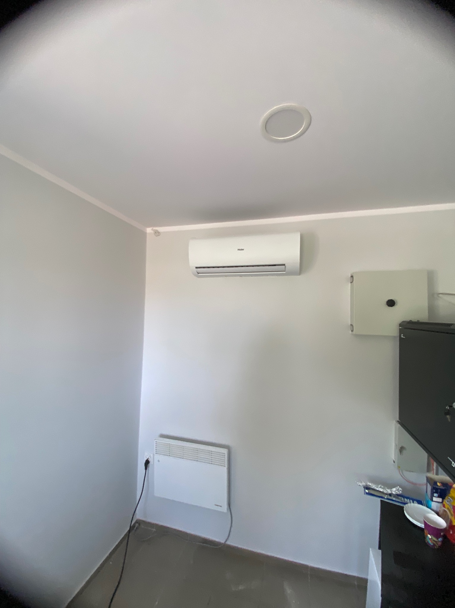 Biała klimatyzacja Haier na ścianie w szarym pomieszczeniu, widoczny grzejnik elektryczny i skrzynka elektryczna. Minimalistyczny design wnętrza.