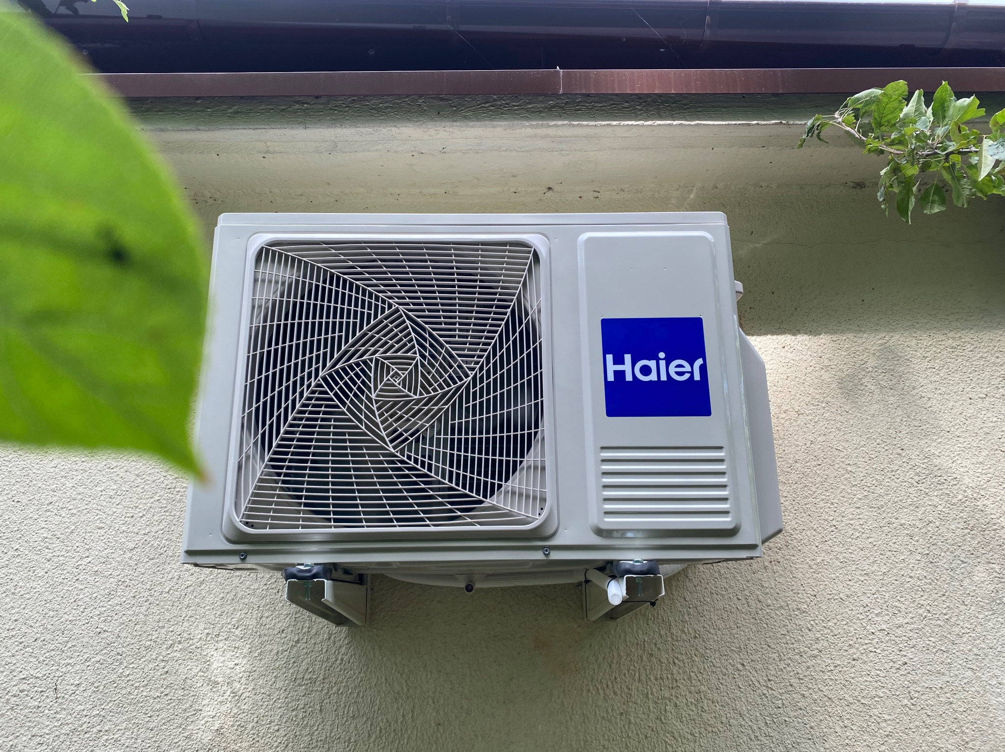 Zamontowana jednostka zewnętrzna klimatyzacji Haier na elewacji budynku, widok z dołu, z fragmentami zieleni w kadrze. Widoczne mocowania i detale konstrukcyjne.