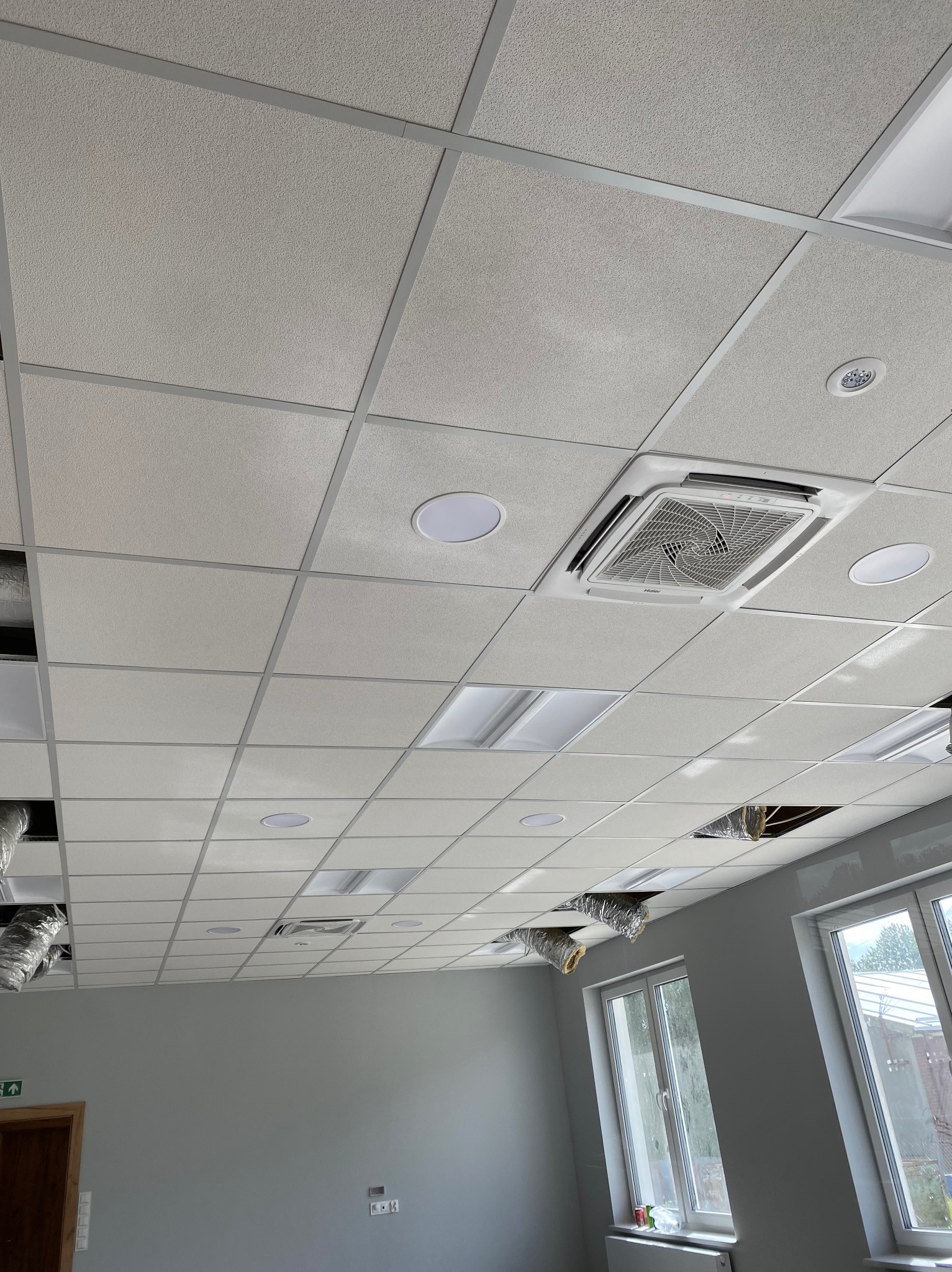 Sufit podwieszany z zamontowaną kasetą klimatyzacyjną Haier i oświetleniem LED. Widoczne otwory na instalacje wentylacyjne i okna z widokiem na zewnątrz.
