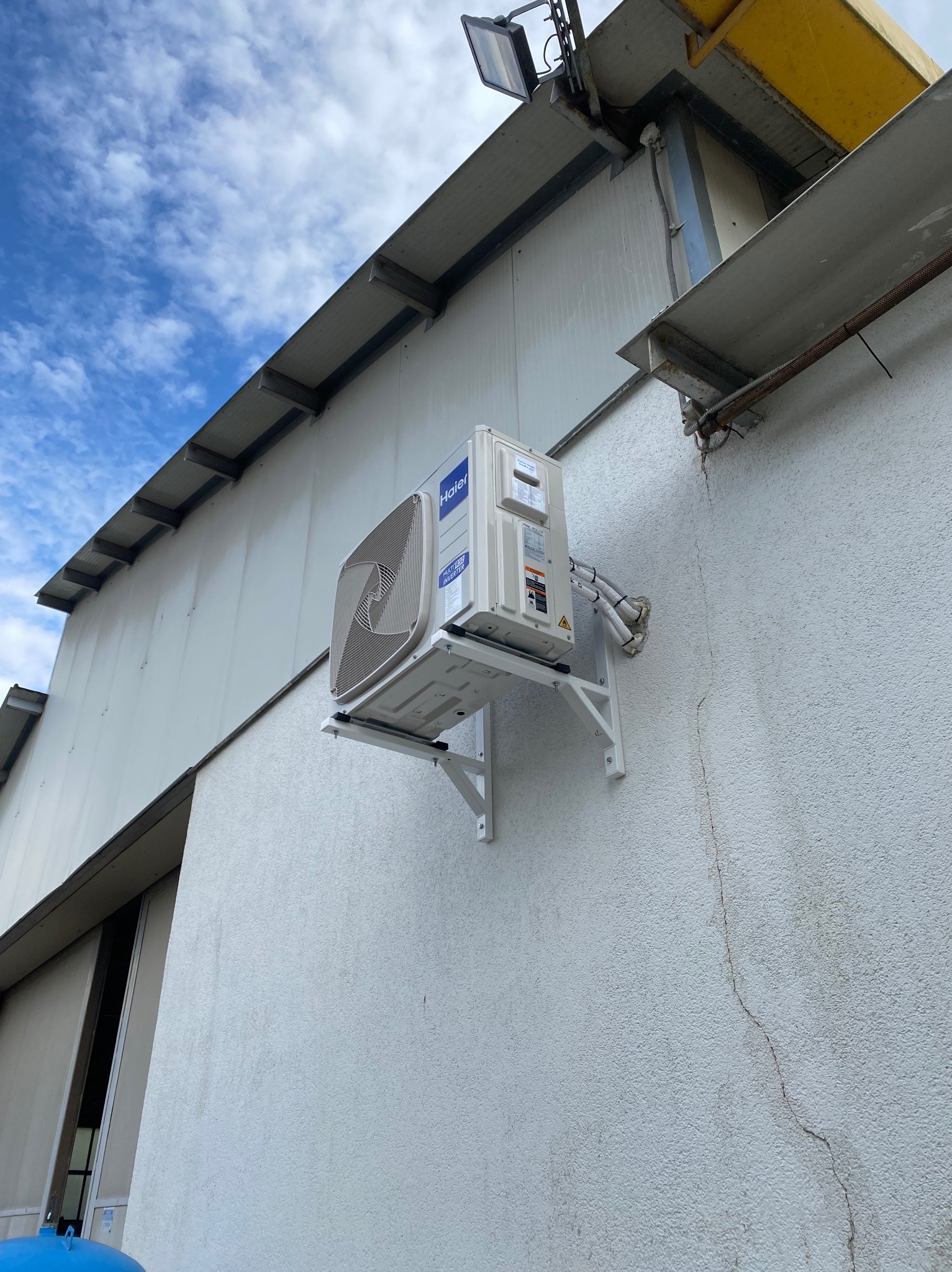 Biały klimatyzator Haier zamontowany na wsporniku na teksturowanej ścianie budynku. Widoczne przewody i elementy instalacji. Ujęcie z dołu, błękitne niebo w tle.