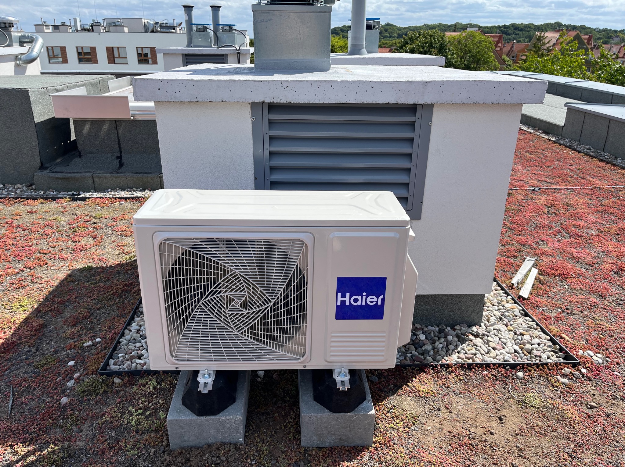 Biała jednostka zewnętrzna klimatyzacji Haier na dachu budynku, zamontowana na betonowych podstawach, z widoczną wentylacją i zielonym dachem.