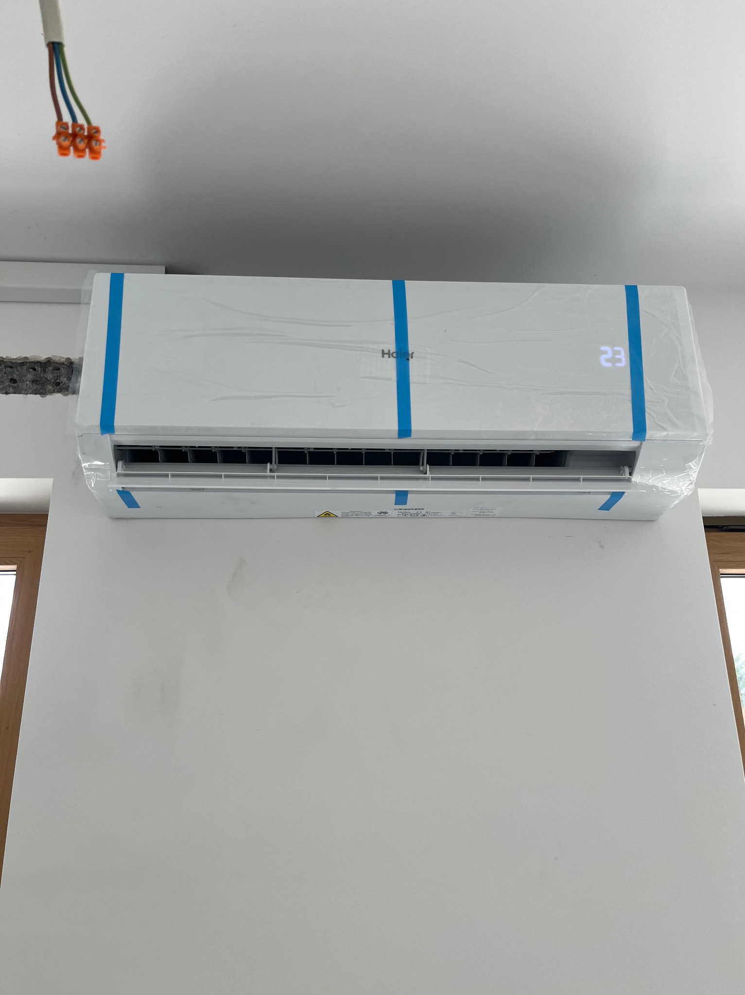 Biały klimatyzator Haier, jeszcze w folii ochronnej z niebieską taśmą, zamontowany na białej ścianie. Widoczne okno i wystające kable.