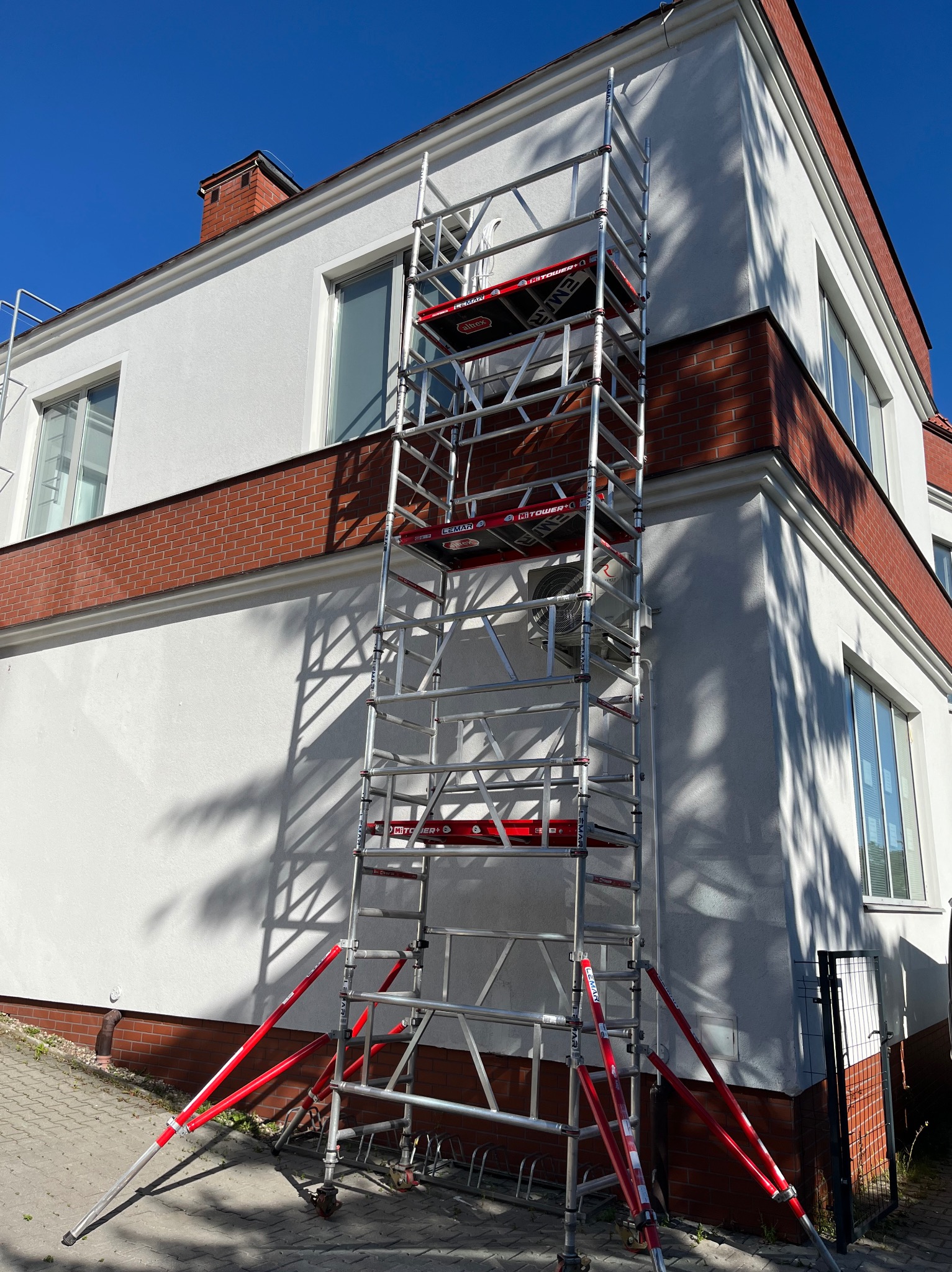 Montaż klimatyzacji na elewacji domu z użyciem rusztowania aluminiowego. Widoczna jednostka zewnętrzna klimatyzatora. Budynek z białymi ścianami i ceglanymi elementami.