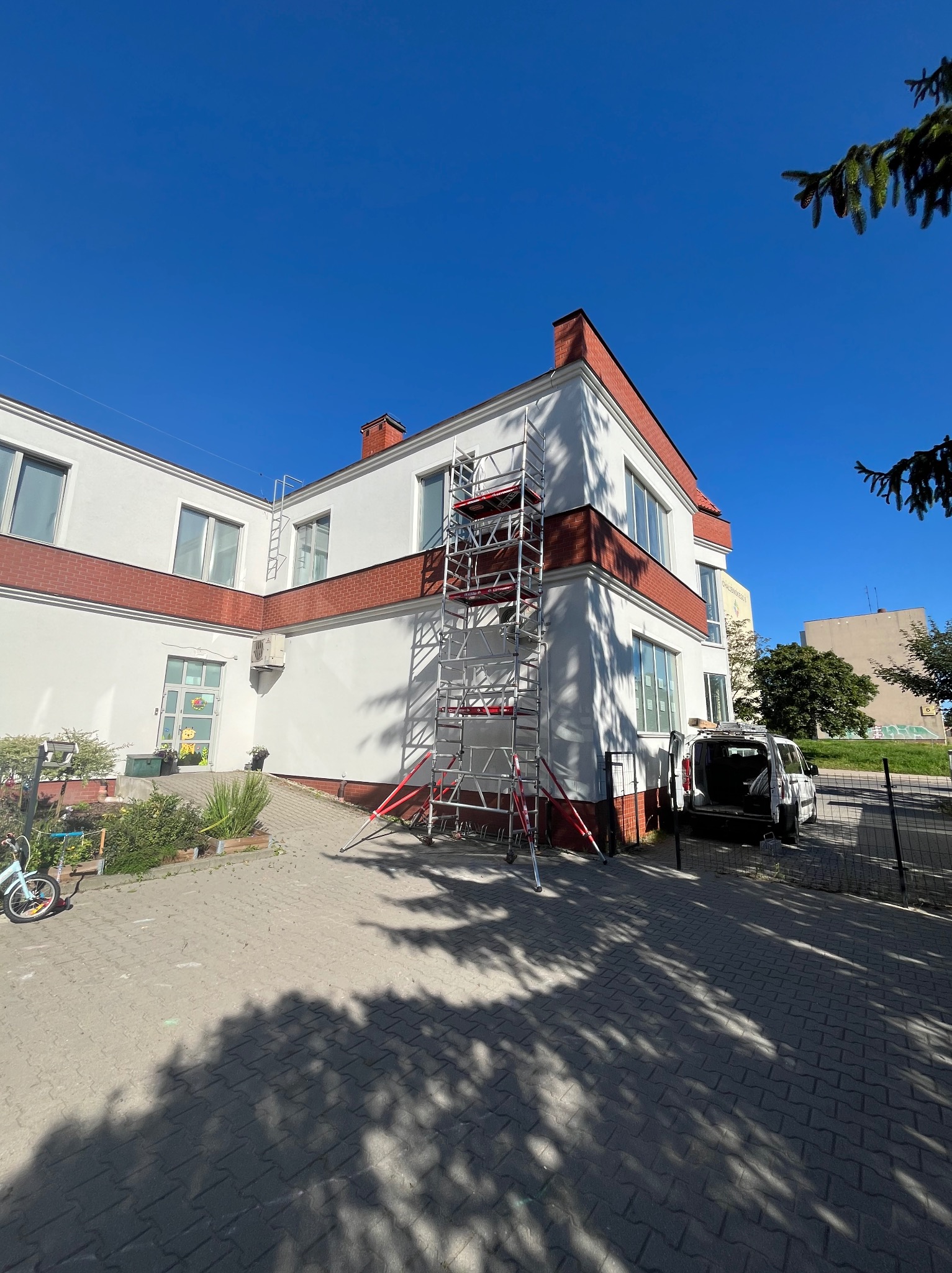 Biały budynek z czerwonymi akcentami, z zamontowaną klimatyzacją i rozstawionym rusztowaniem. Widoczny samochód dostawczy i błękitne niebo.