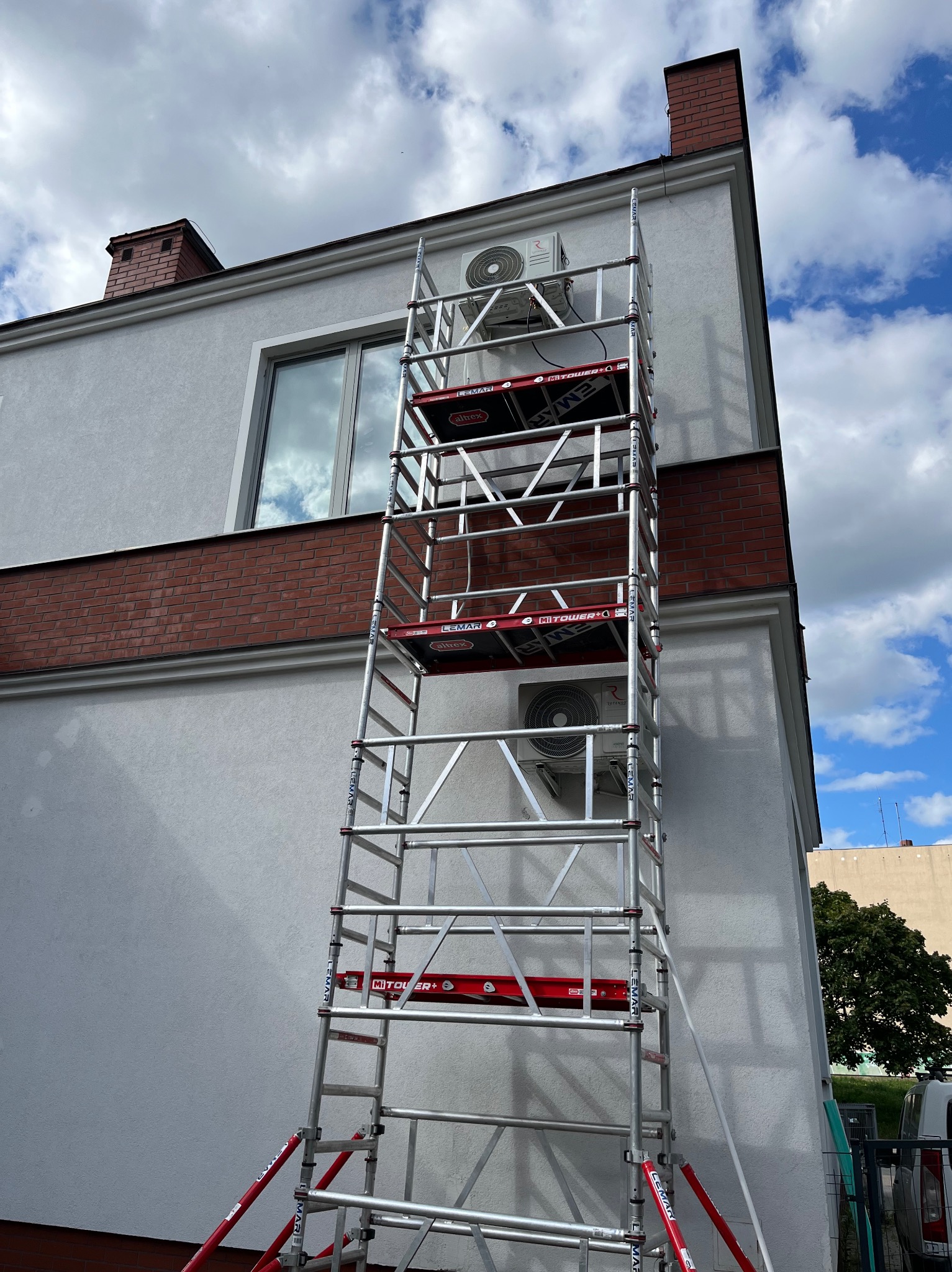 Montaż klimatyzacji na elewacji domu z użyciem rusztowania. Widoczne jednostki zewnętrzne klimatyzatorów. Biała elewacja z ceglanym pasem i kominem.