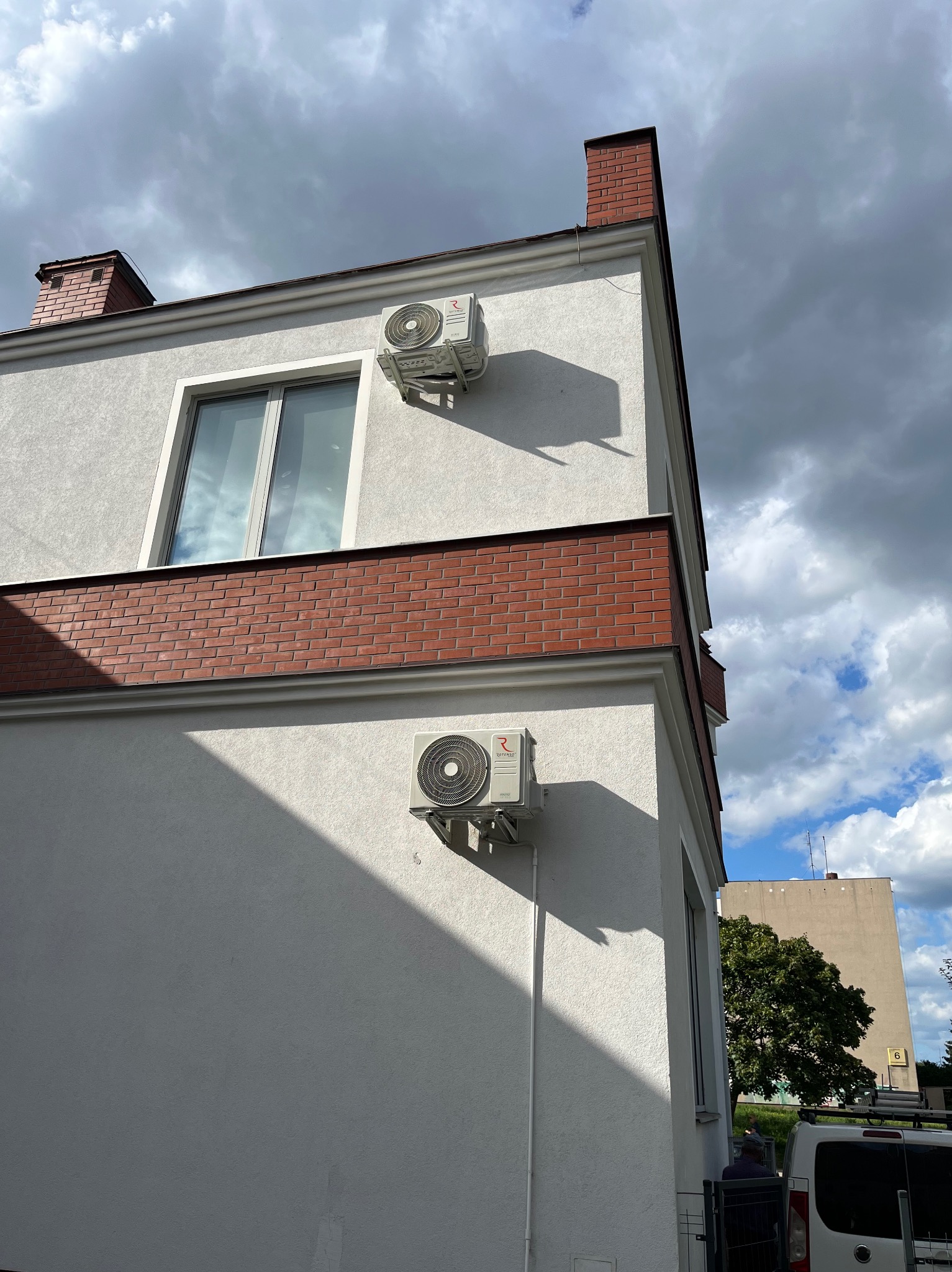 Dwa klimatyzatory na elewacji budynku z ceglanymi elementami dekoracyjnymi, widoczne z dołu na tle pochmurnego nieba. Jeden klimatyzator zamontowany nad oknem.