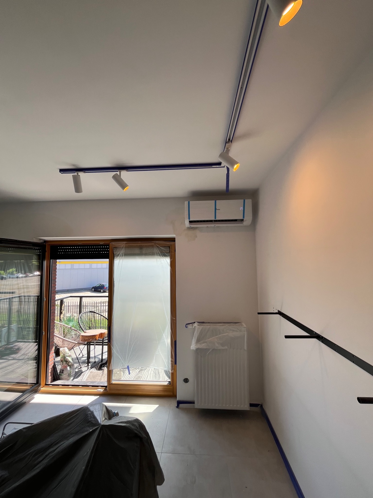 Biały klimatyzator na ścianie w jasnym pokoju z niebieskimi akcentami, widoczne szyny oświetleniowe i zabezpieczone okno folią malarską.