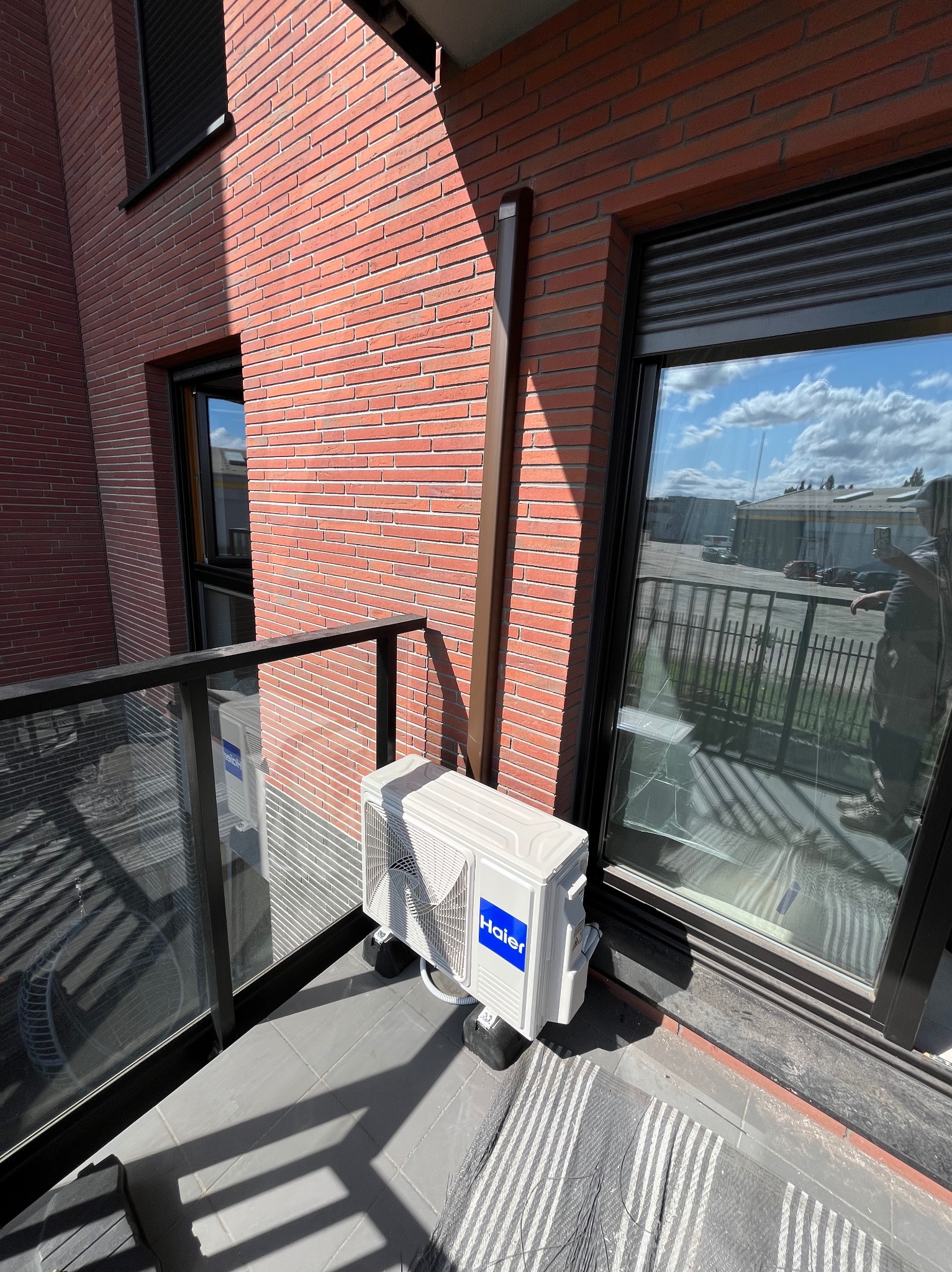 Biała jednostka zewnętrzna klimatyzacji Haier zamontowana na balkonie, widoczna na tle ceglanej ściany budynku i okna z roletami. Widok z dołu, z boku.