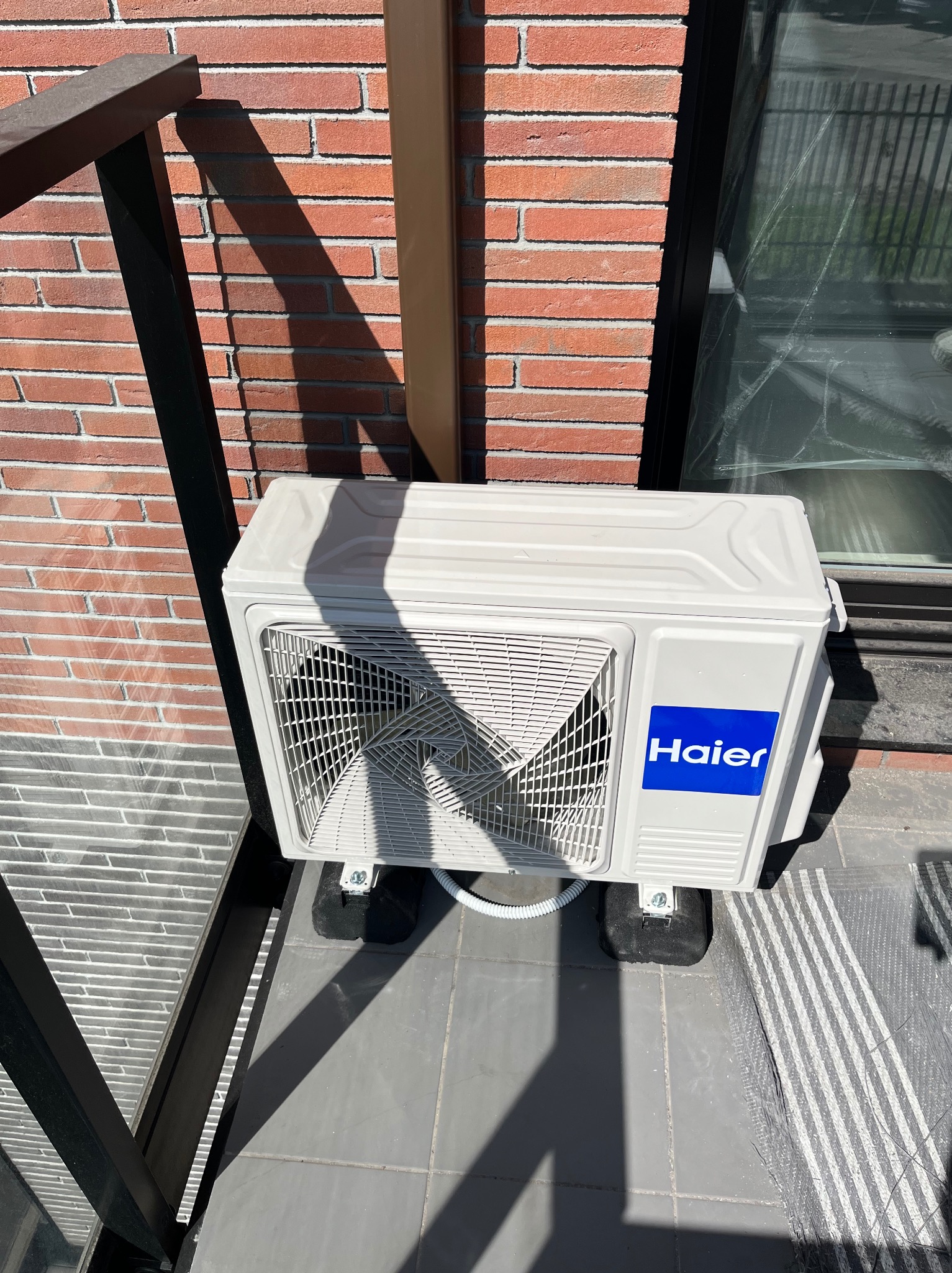 Biała jednostka zewnętrzna klimatyzacji Haier zamontowana na szarym balkonie, z widoczną ceglaną ścianą i fragmentem okna w tle, w słoneczny dzień.