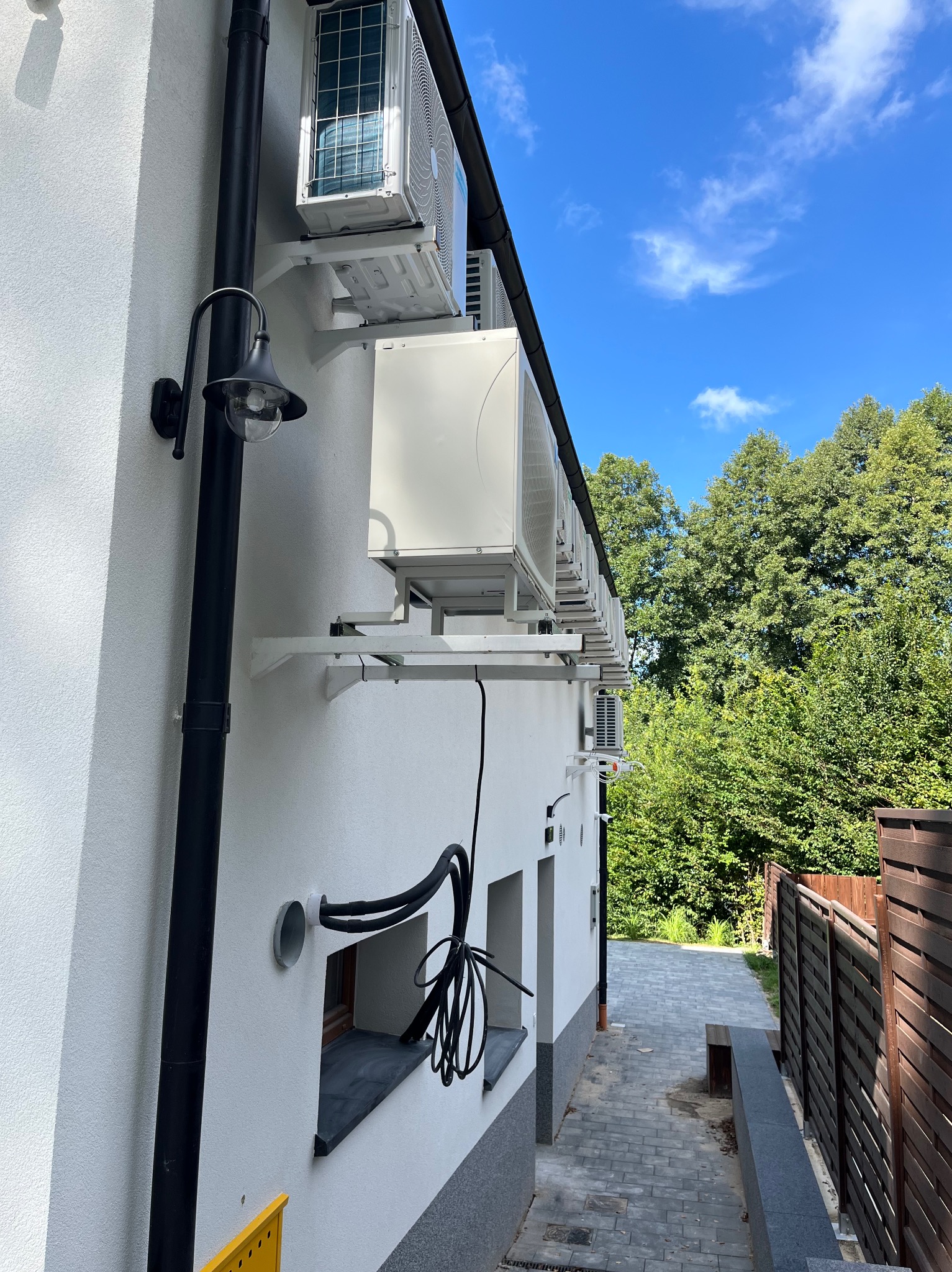 Biała ściana budynku z zamontowanymi jednostkami klimatyzacji, widoczne przewody i lampa zewnętrzna. Błękitne niebo i zieleń drzew w tle.