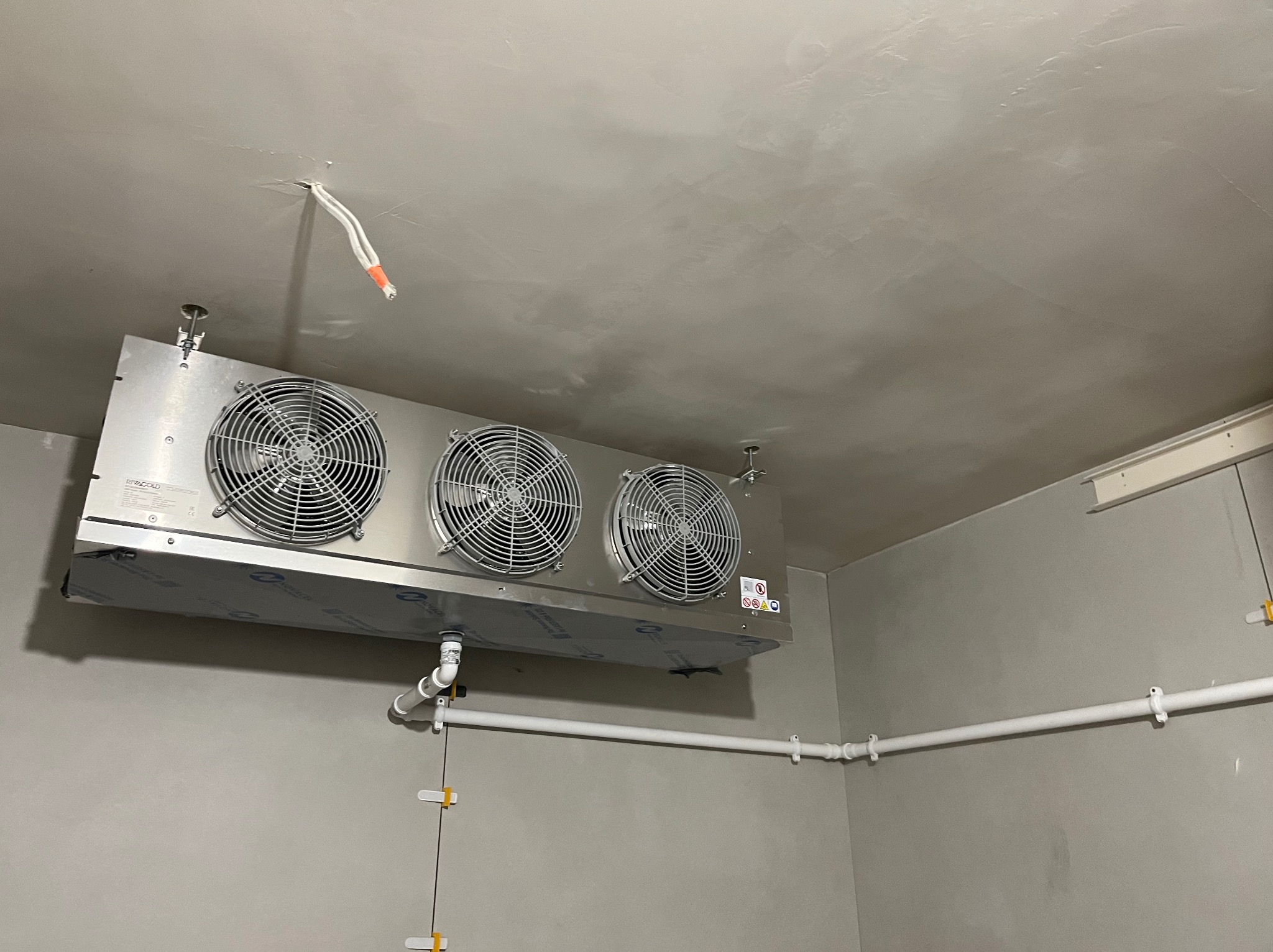 Zamontowany, przemysłowy klimatyzator sufitowy z trzema wentylatorami w szarej przestrzeni. Widoczne rury i kable. Chłodne oświetlenie.