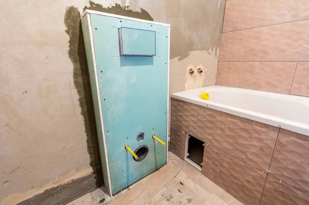 Montaż stelaża podtynkowego z płyt gipsowo-kartonowych w łazience, widoczne rury doprowadzające wodę, obok wanna z obudową z płytek ceramicznych w trakcie wykańczania.