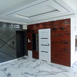 Usługi Remontowo-Budowlane - Nowoczesne wnętrze: grafitowa ściana z geometrycznym wzorem, ceglana ściana z białymi drzwiami i fragment białej kuchni z drewnianym blatem. Marmurowe płytki na podłodze.