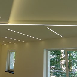 Usługi Remontowo-Budowlane - Nowoczesne oświetlenie sufitowe z liniowymi lampami LED w jasnym wnętrzu. Widoczne okna z widokiem na zieleń. Minimalistyczny design, świeżo po remoncie.