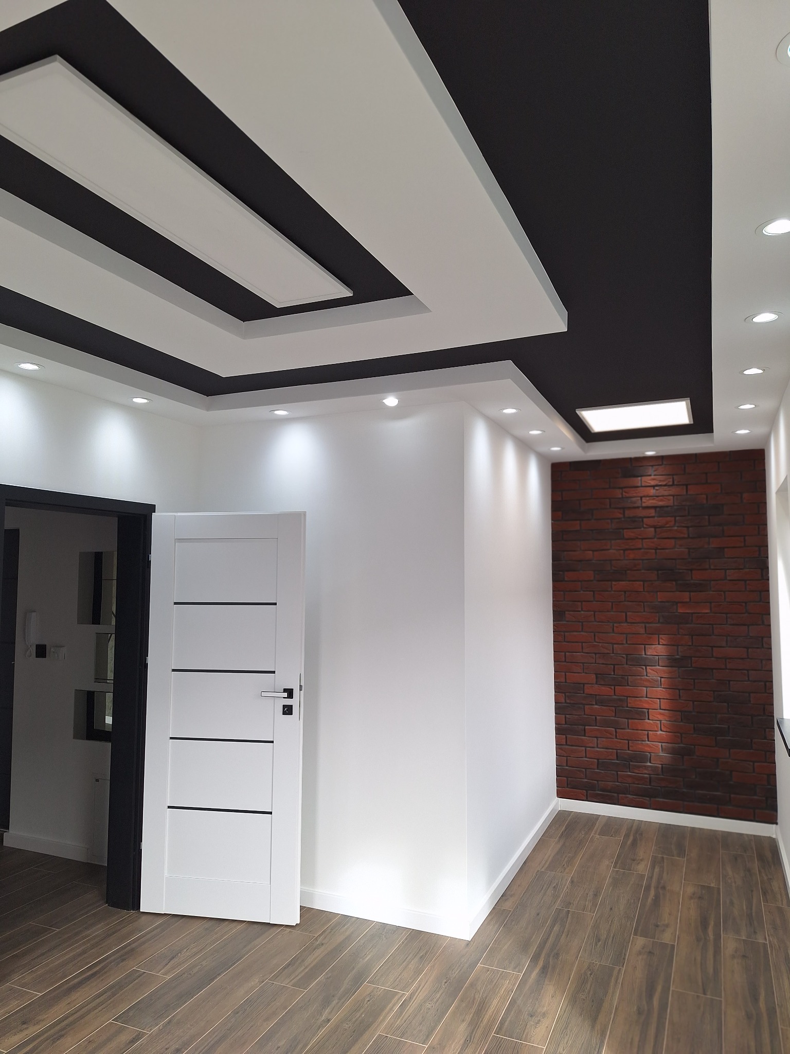 Nowoczesne wnętrze po remoncie: białe ściany, drewnopodobna podłoga, ceglana ściana akcentująca i designerski sufit z oświetleniem. Białe drzwi z czarnymi akcentami.