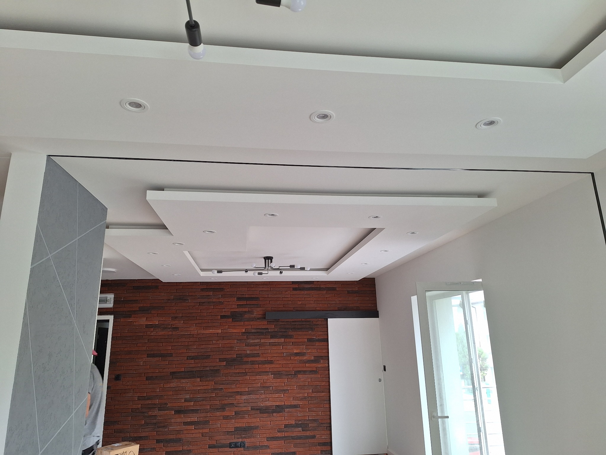 Nowoczesne wykończenie wnętrza: ceglana ściana, podwieszany sufit z oświetleniem punktowym i dekoracyjna ściana z szarym wzorem geometrycznym.