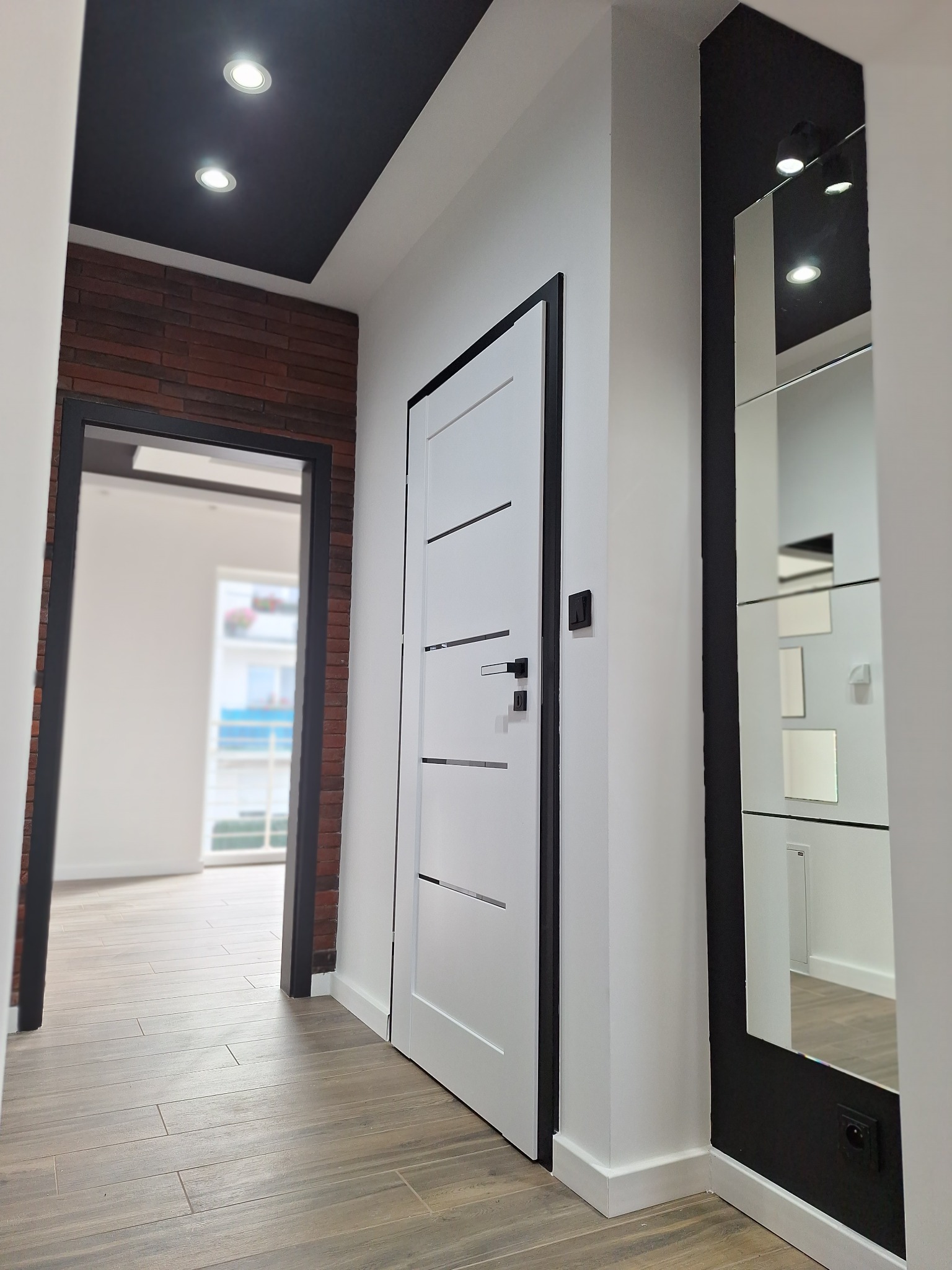 Nowoczesny przedpokój z białą, minimalistyczną stolarką drzwiową, lustrem w czarnej ramie i akcentem ceglanej ściany. Podłoga wyłożona płytkami imitującymi drewno.