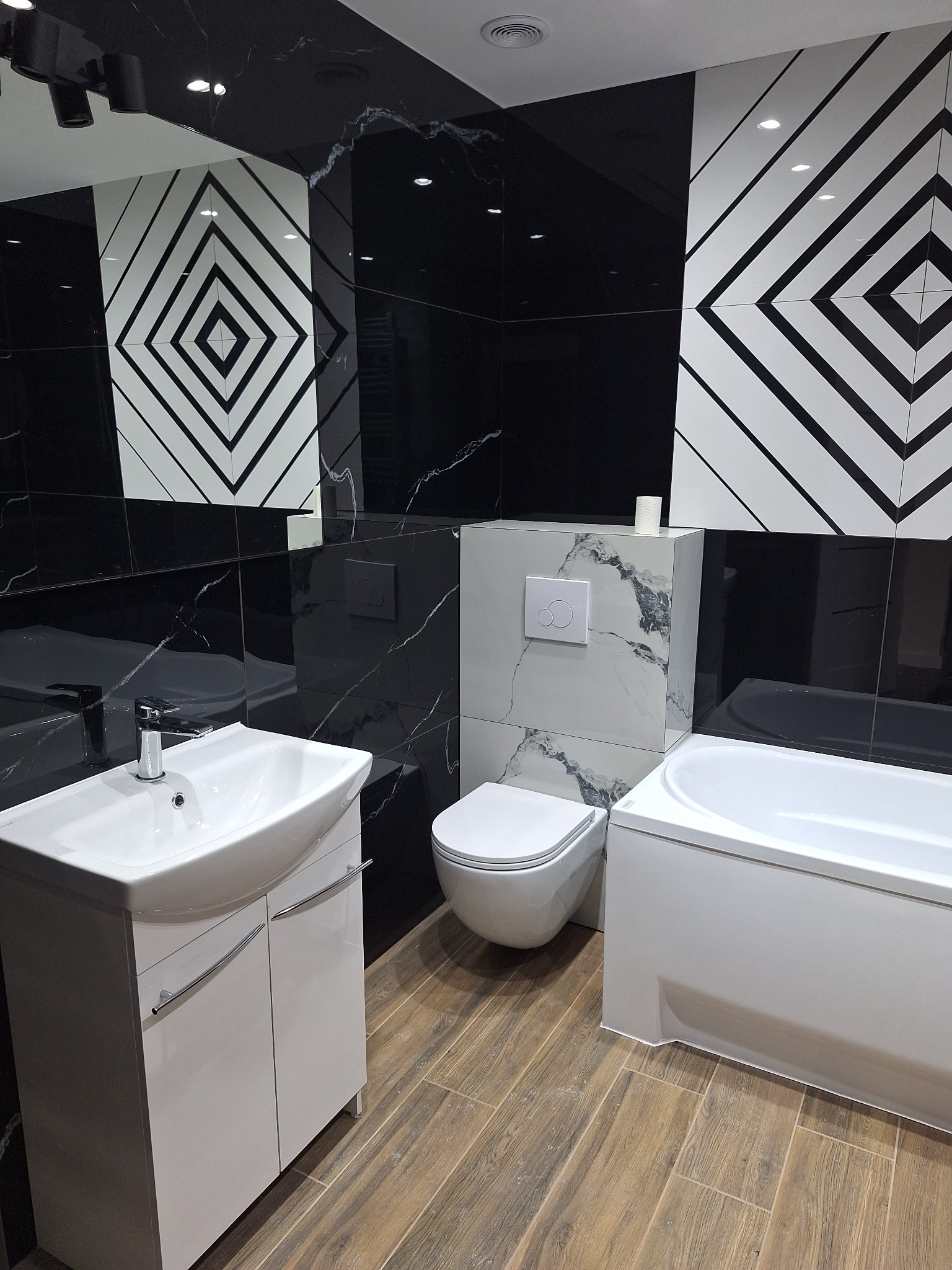 Nowoczesna łazienka z czarno-białymi płytkami w geometryczne wzory, białą armaturą i drewnopodobną podłogą. Umywalka z szafką, toaleta i wanna w jednym kadrze.