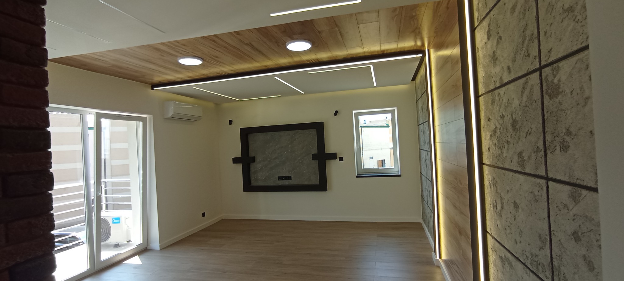 Nowoczesne wnętrze po remoncie: drewniany sufit z oświetleniem LED, ściana z betonowych paneli, okno i drzwi balkonowe. Minimalistyczny design, jasne kolory.