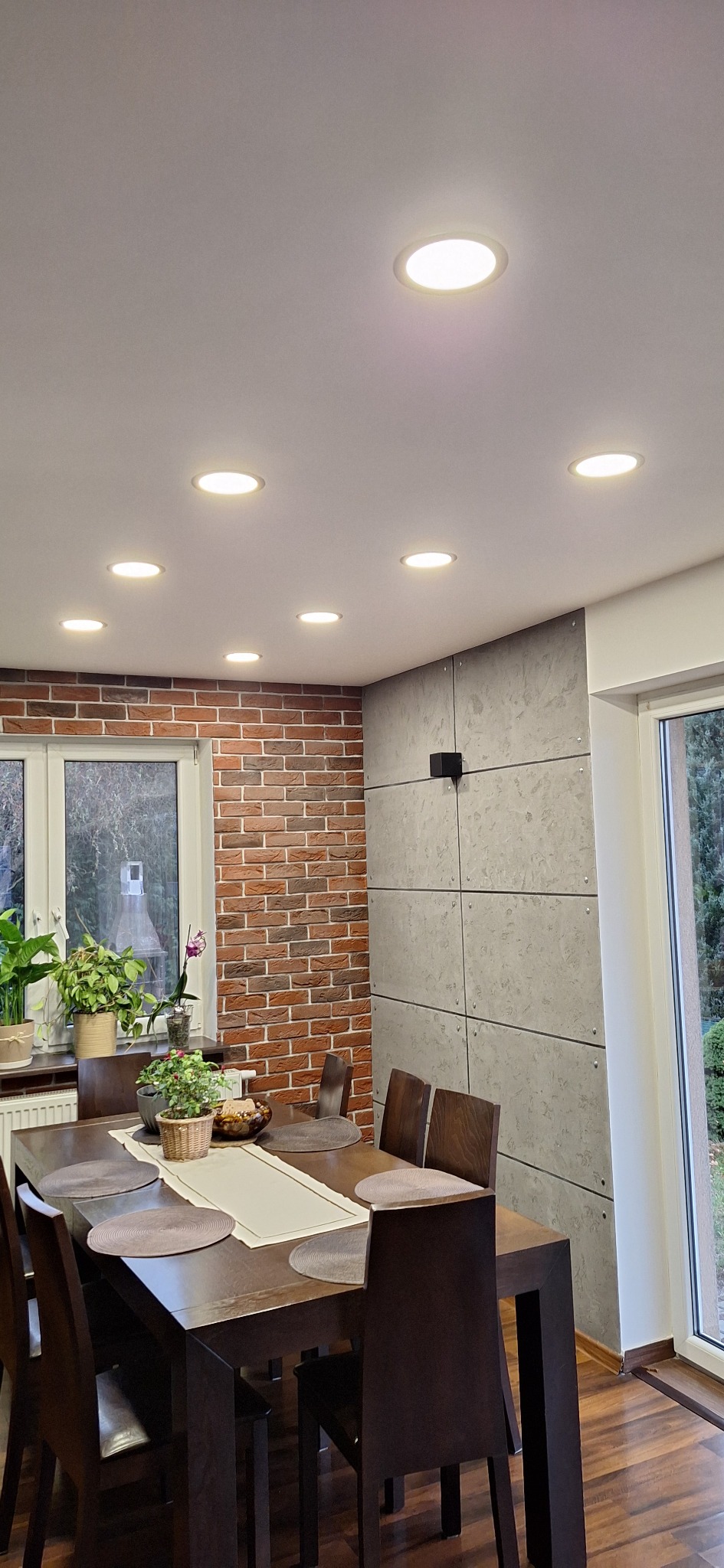 Jadalnia po remoncie: ceglana ściana, betonowe panele, drewniany stół z dekoracjami i oświetlenie sufitowe. Widok na okno i ogród.