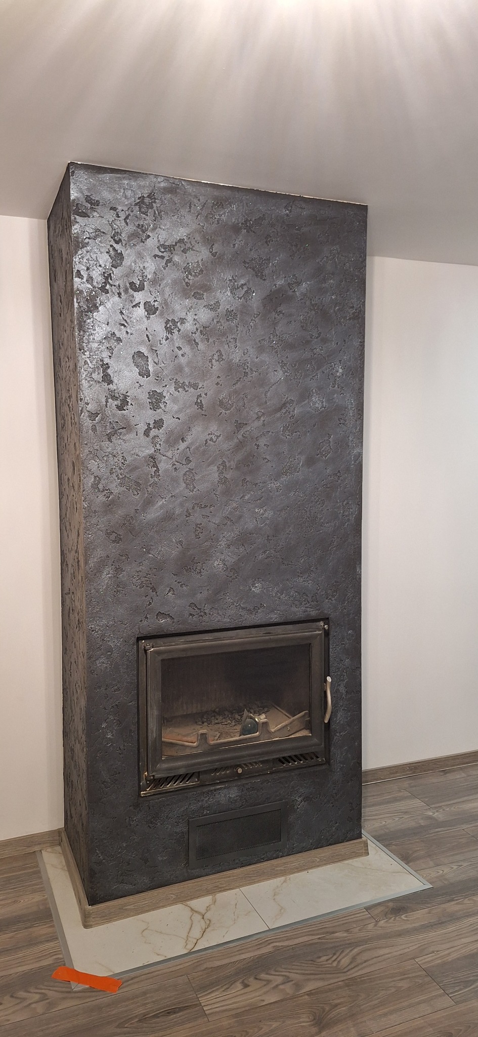 Otynkowany kominek w ciemnych barwach z metalowymi drzwiczkami, wykończony płytkami imitującymi marmur. Podłoga z paneli w odcieniach brązu.