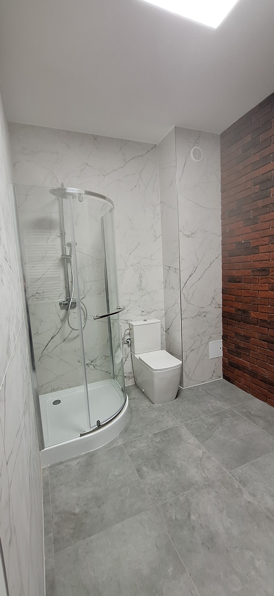 Nowoczesna łazienka po remoncie: kabina prysznicowa z zaokrąglonymi drzwiami, toaleta i ściana z imitacji cegły. Szare płytki podłogowe i marmurowe ściany.
