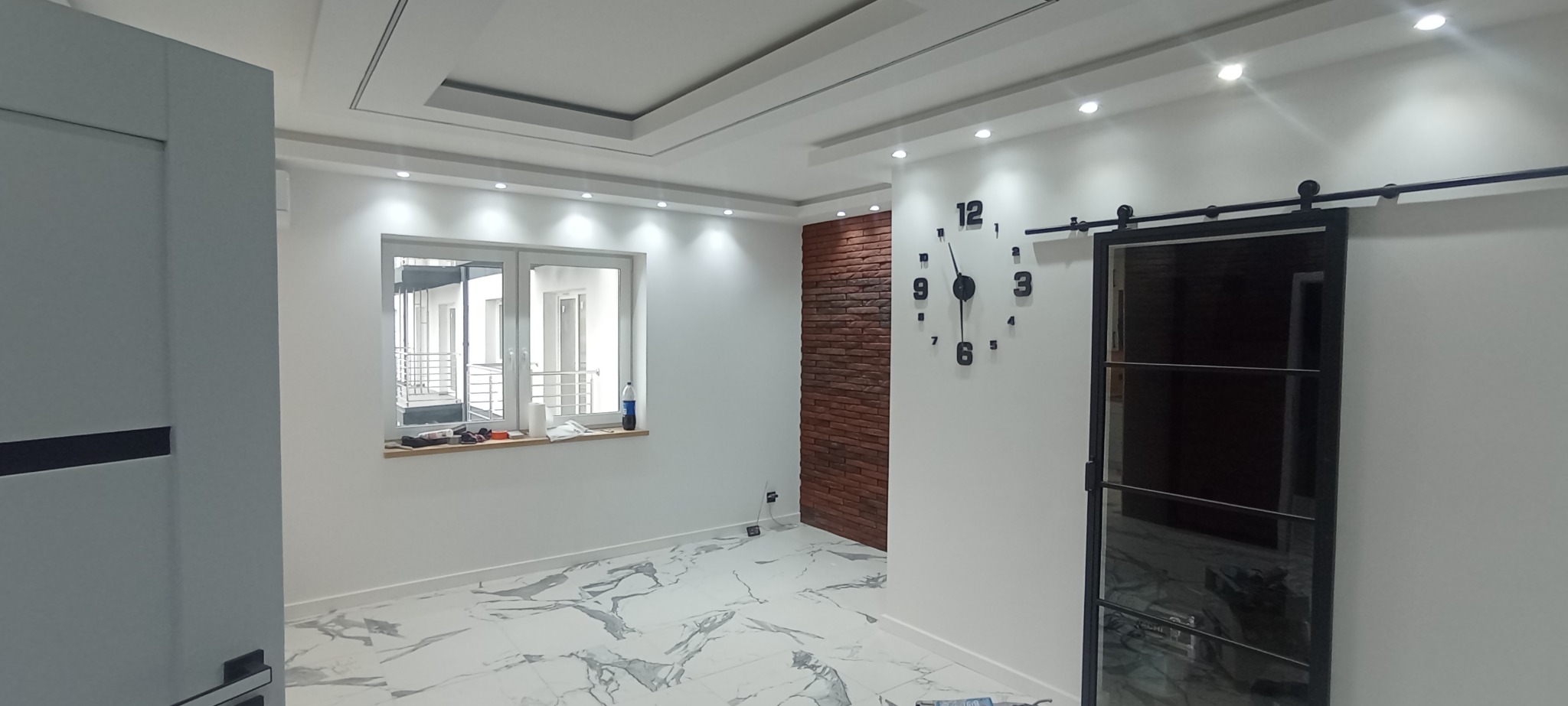 Nowoczesne wnętrze po remoncie: białe ściany, marmurowe płytki, ceglana ściana akcentowa, przesuwne drzwi stodołowe i duży zegar ścienny.